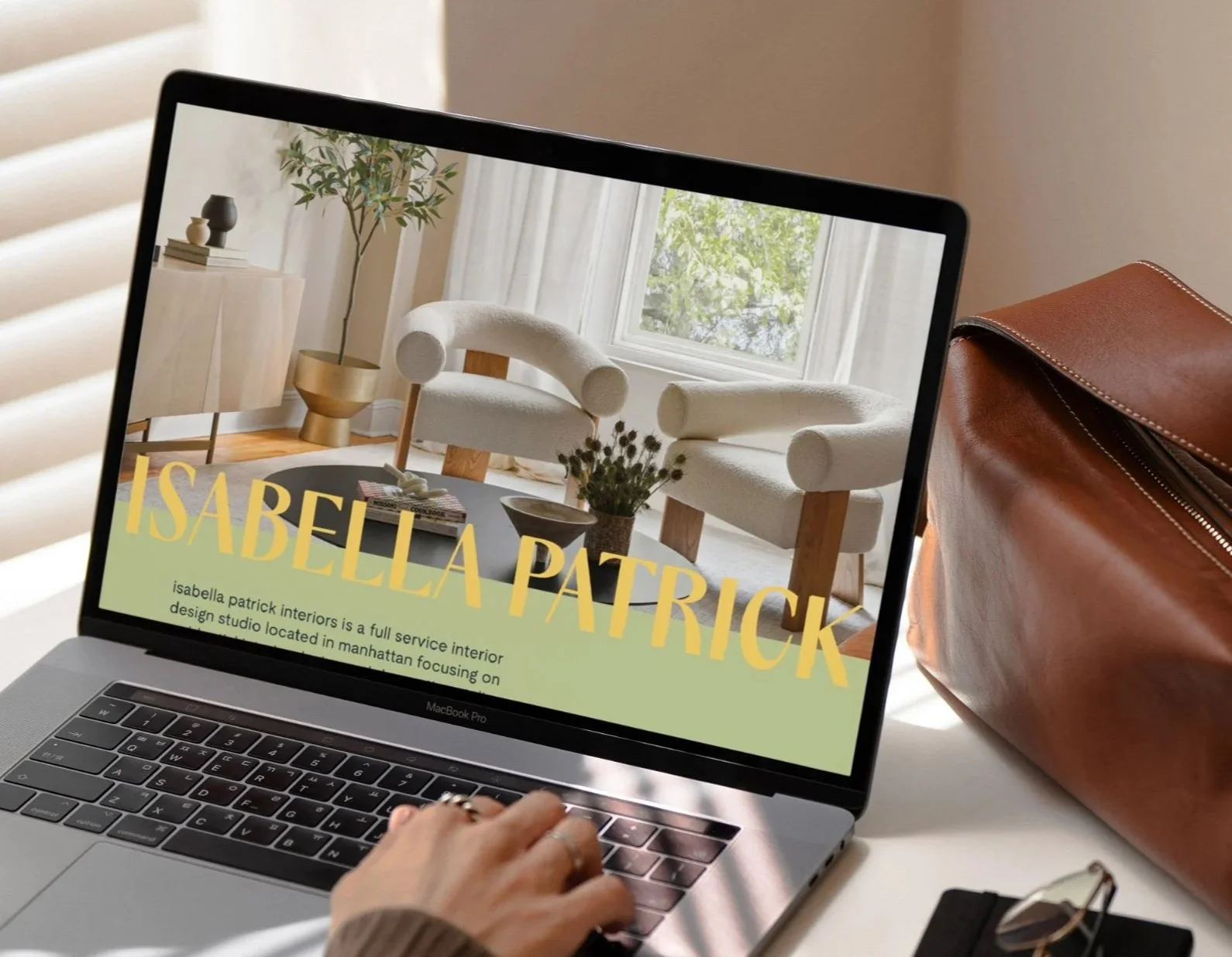 A person using a MacBook Pro laptop on a white table, with a photo of a modern interior design living room on the screen, featuring a beige sofa, a round coffee table with books and a plant, and a window with natural light. There is a brown leather bag next to the laptop.