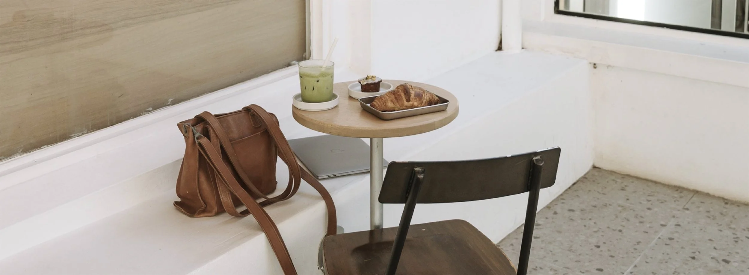 Small round table with a green iced drink, a chocolate muffin, and a croissant in a metal tray, next to a black chair and a brown handbag outside a window.