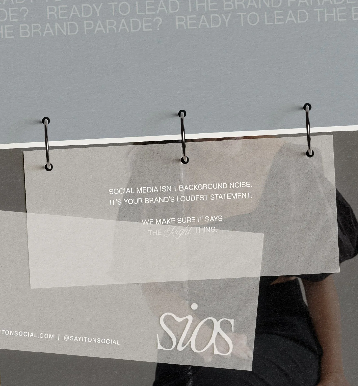 A close-up of a hanging paper with a printed message, attached by metal rings. The message says, "Social media isn't background noise. It's your brand's loudest statement. We make sure it says the right thing." The background includes faint printed text and partial view of a person.