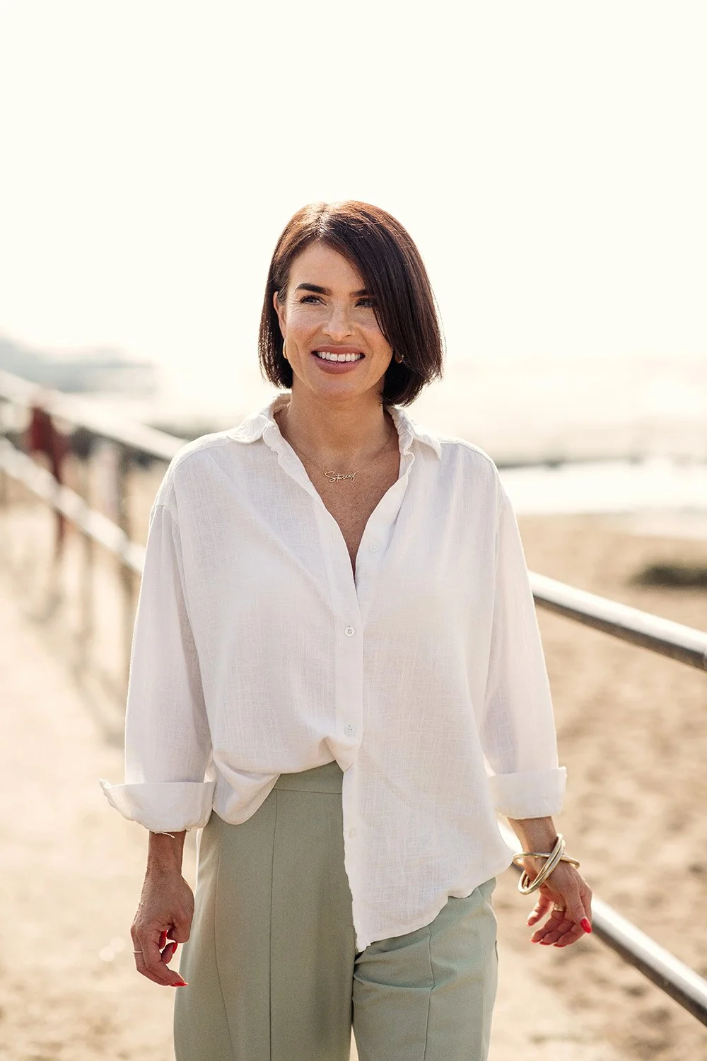 A woman standing outdoors near the beach wearing a white shirt and light-colored trousers, smiling with the ocean and a walkway in the background.