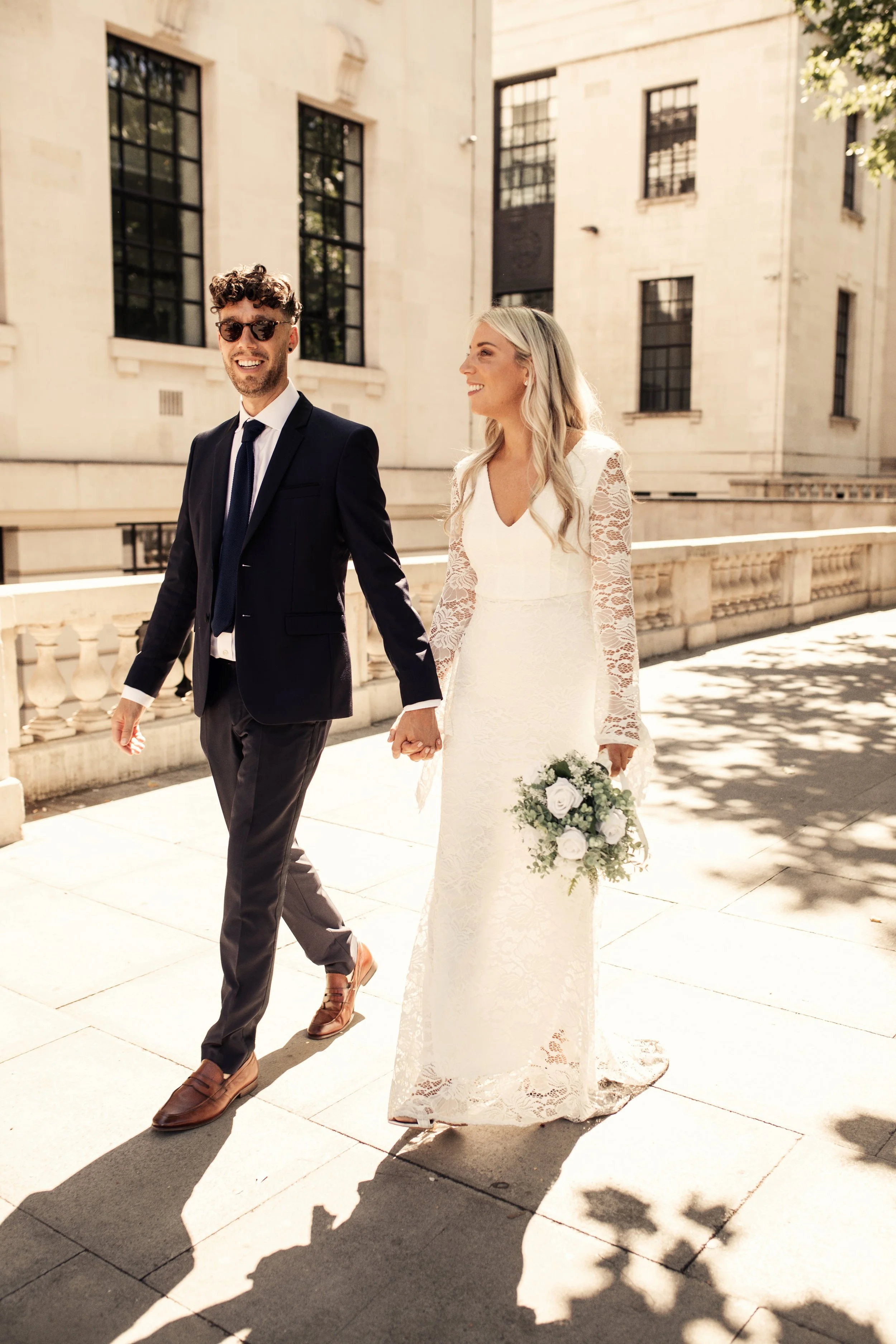 Bride and groom walking in the sunshine, hand in hand, outside Marylebone Town Hall after their wedding.