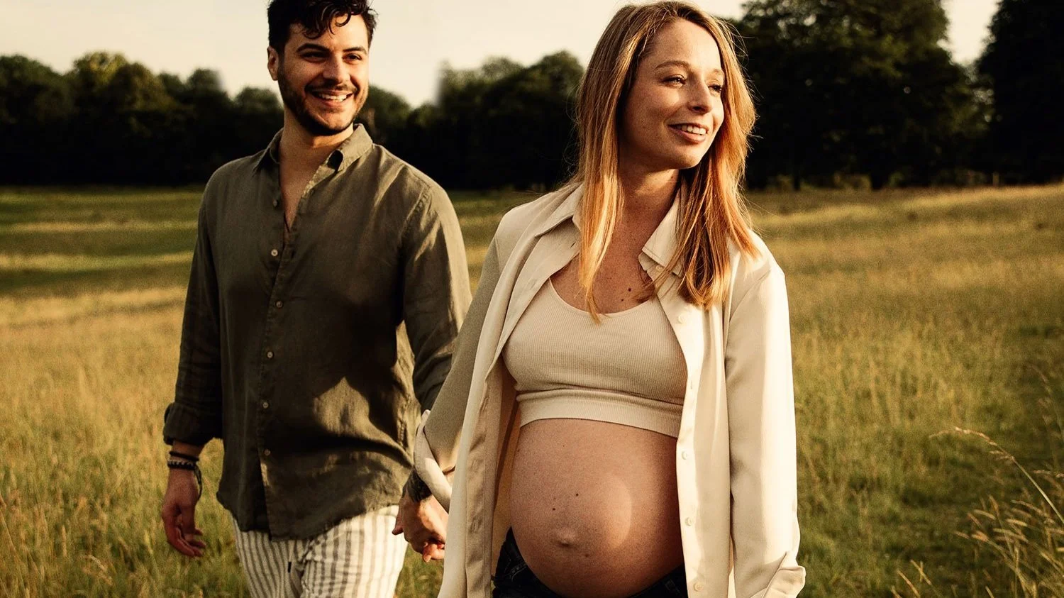 Golden hour in a field. A pregnant woman looks off in the distance, holding hands with her partner who is looking happily at her.