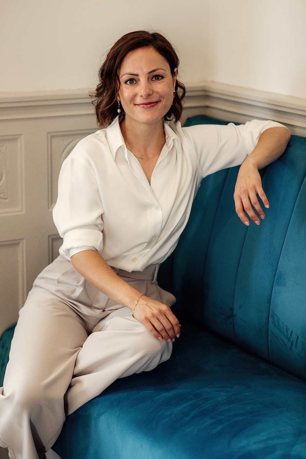 A woman in a white blouse and beige trousers sitting on a blue sofa, smiling at the camera, in a room with beige walls