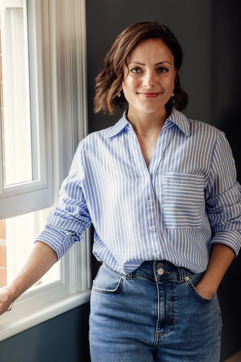 A woman with short wavy brown hair, smiling, wearing a blue and white striped button-up shirt with rolled-up sleeves and blue jeans, standing by a window with natural light.