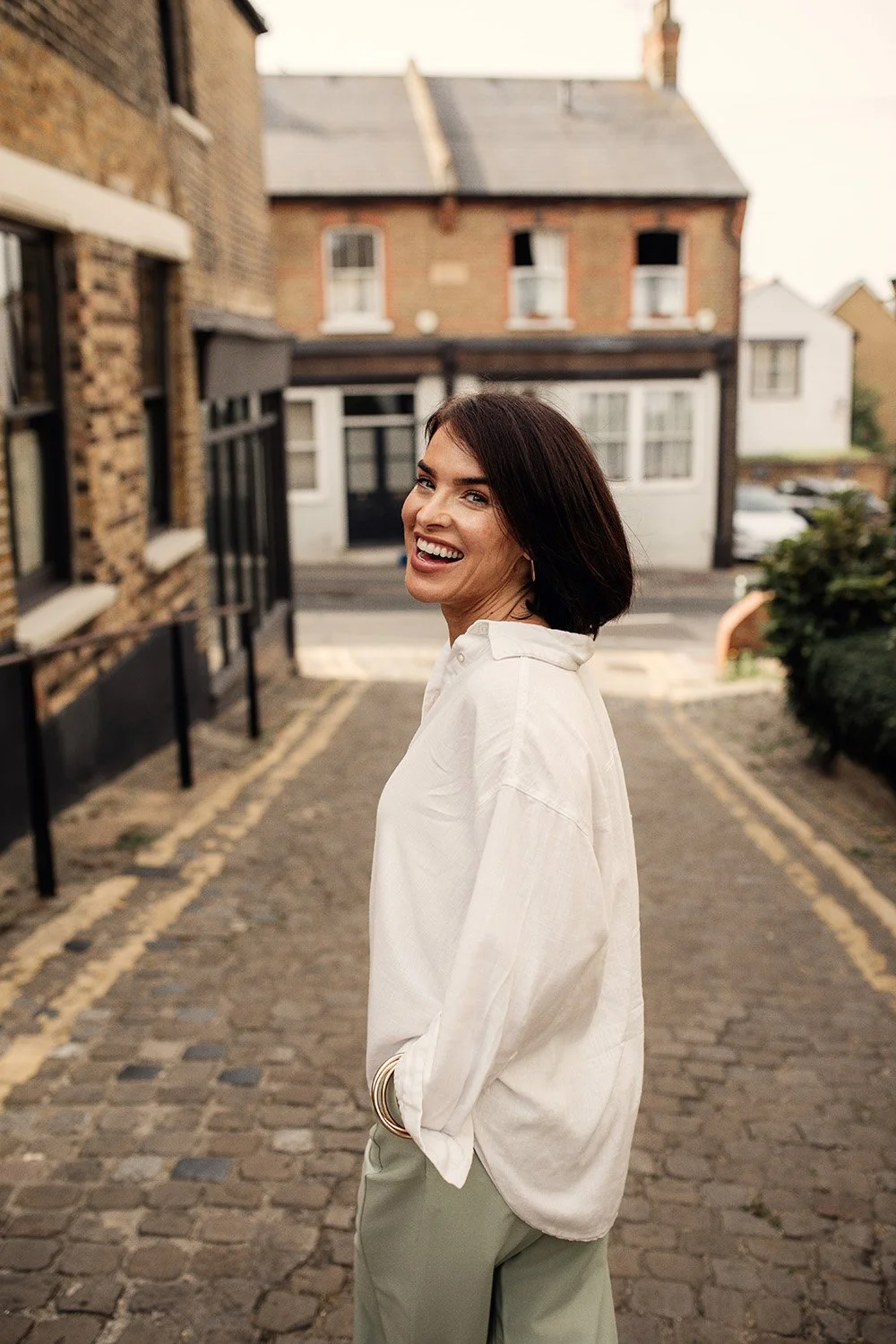 A woman with dark hair, smiling and looking over her shoulder, standing on a cobblestone street in front of old brick and white houses.