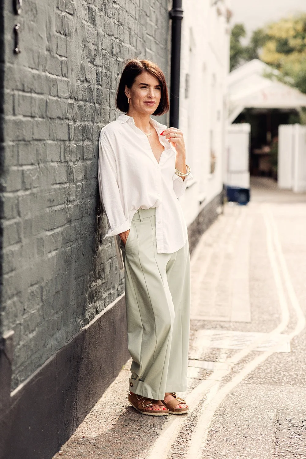 Woman standing against a brick wall wearing a white shirt and light-colored wide-leg trousers, outdoors on a city street.