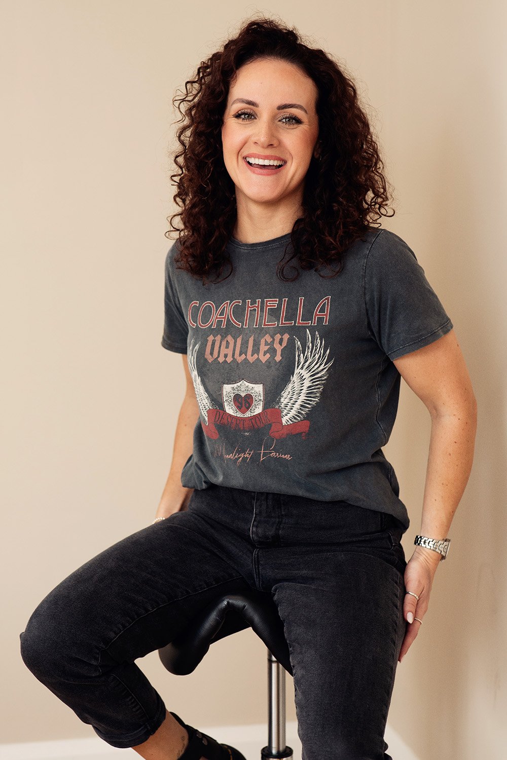 A woman with curly brunette hair sitting on a stool, wearing a dark gray 'Coachella Valley' t-shirt and black jeans, smiling at the camera.