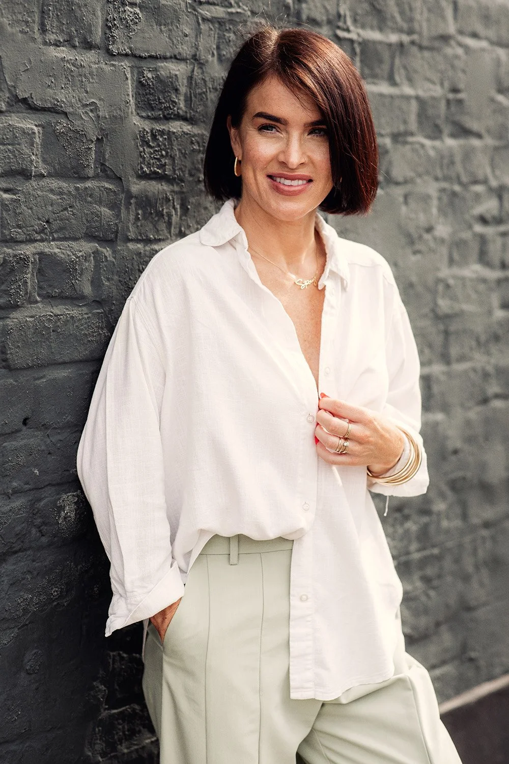 A woman with short brown hair wearing a white button-down shirt and light-colored trousers, leaning against a dark gray brick wall, smiling at the camera.