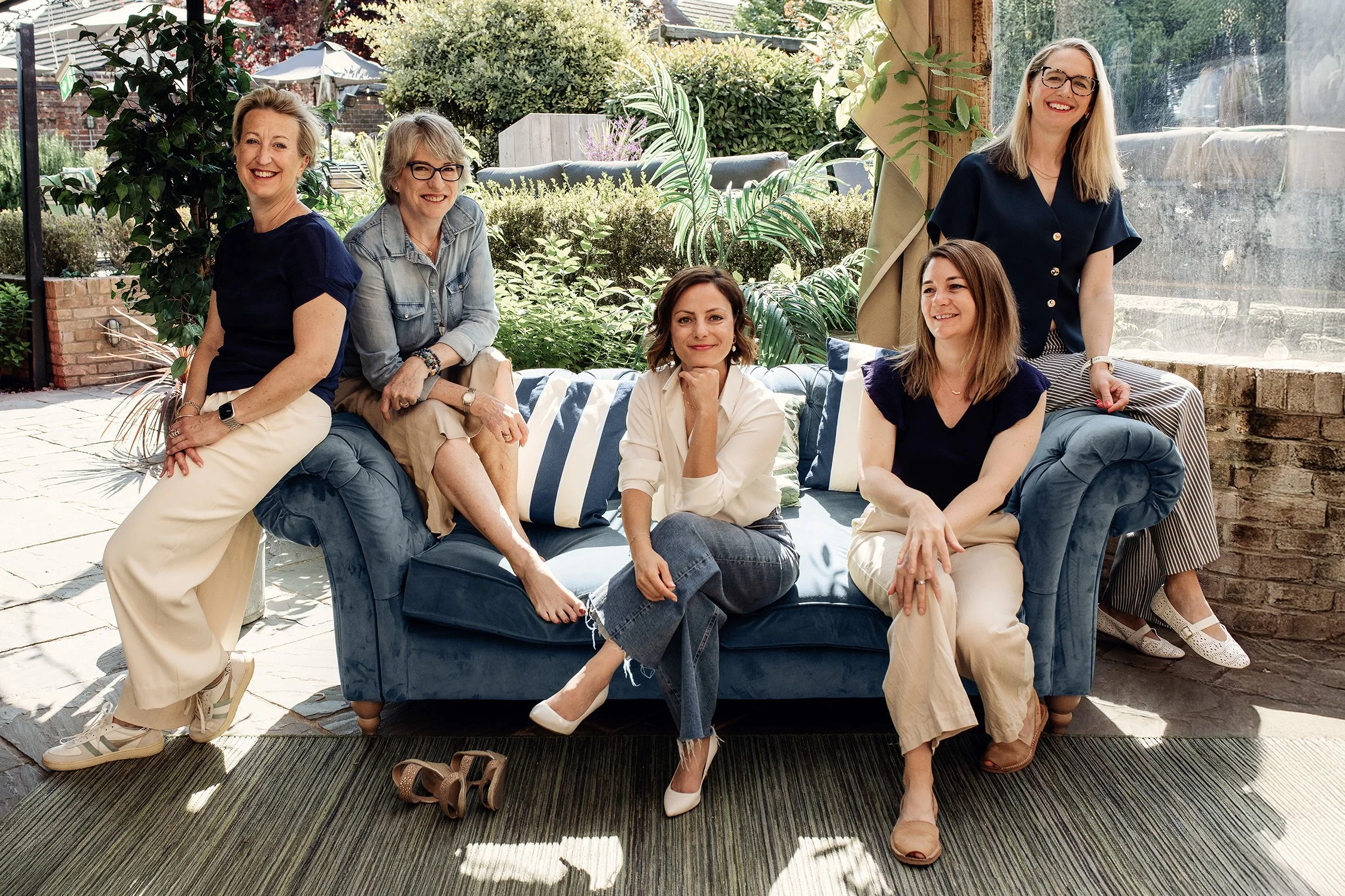 Five women sitting and standing around a blue velvet couch in a sunlit garden patio with greenery and brick walls.