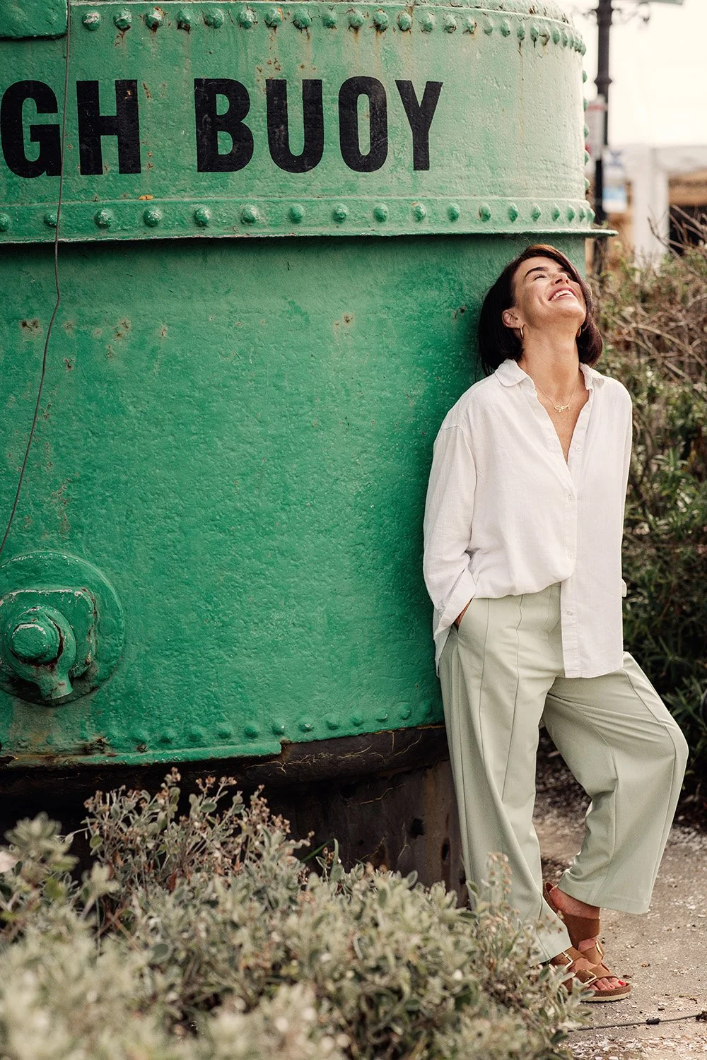 A woman leaning against a large green structure marked 'LEIGH BUOY', smiling with her eyes closed laughing, hands in pockets, wearing a white shirt and beige trousers, outdoors with plants around.