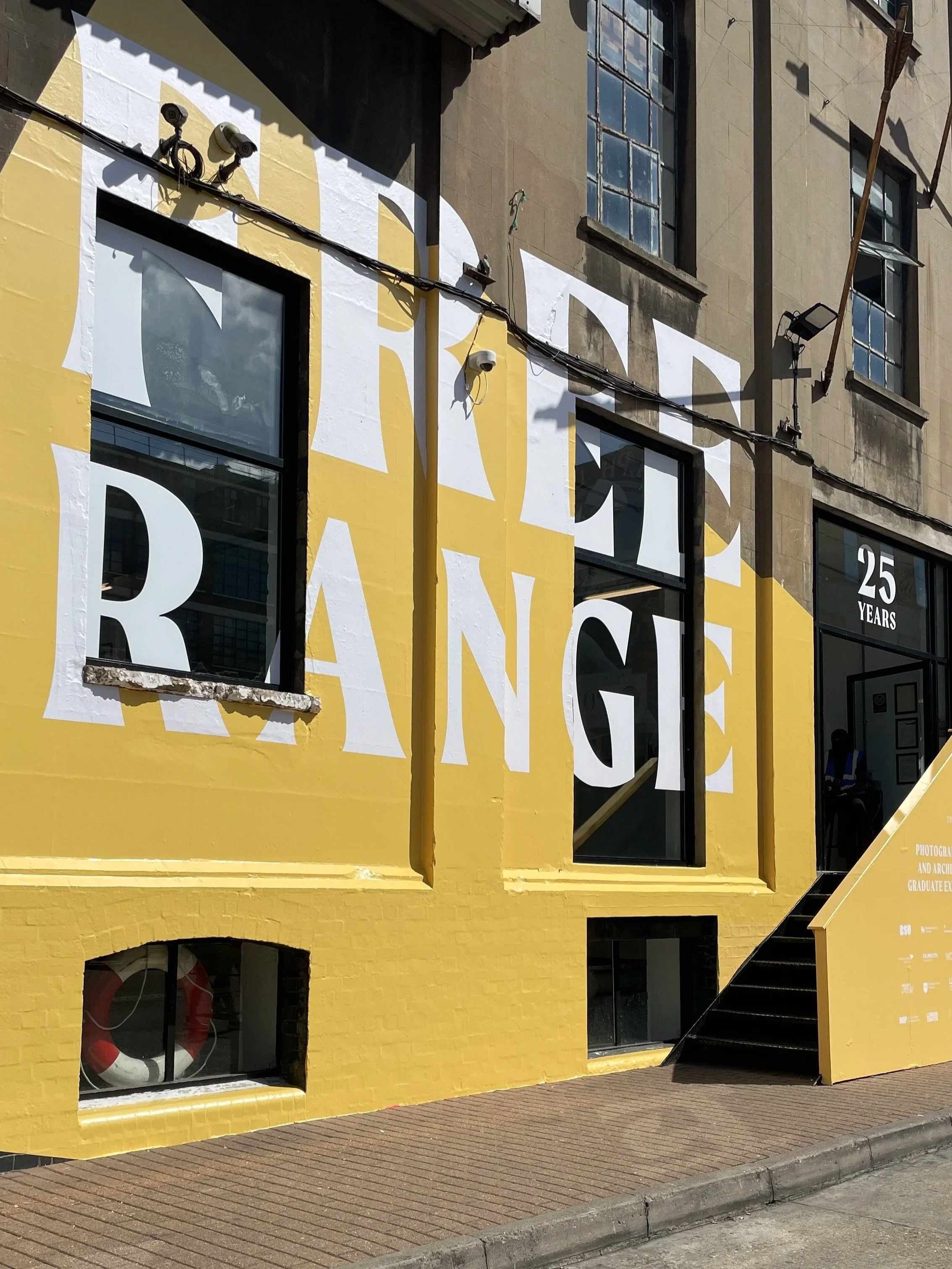Yellow-painted building with large white letters spelling 'THE RANG' partly obscured, and a window in the yellow wall. The building has a small set of black stairs leading to an entrance, with a partial view of a person inside.