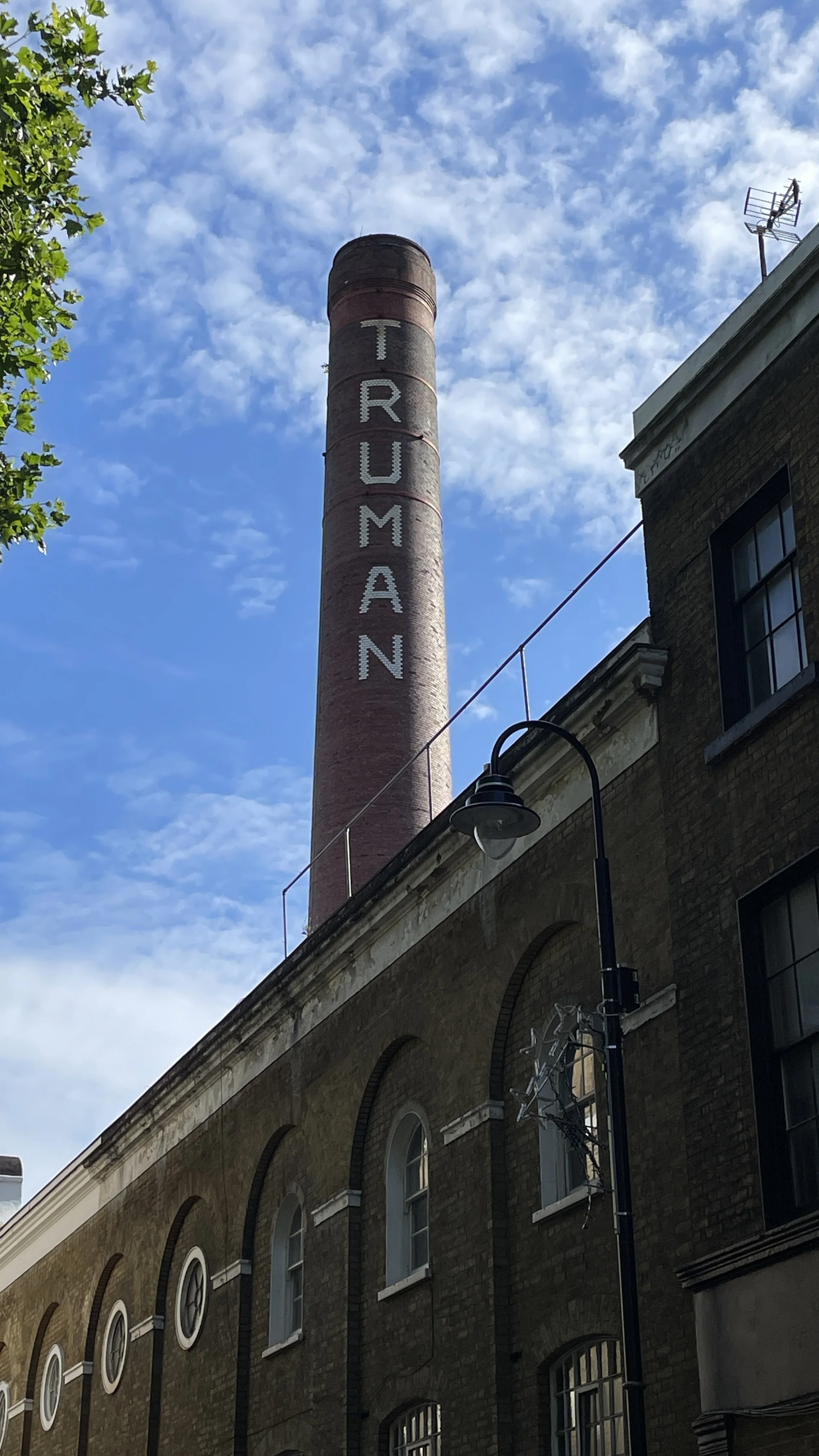 A tall brick chimney with the word 'TRUMAN' written vertically on it, set against a background of blue sky with scattered clouds, surrounded by buildings and streetlights.