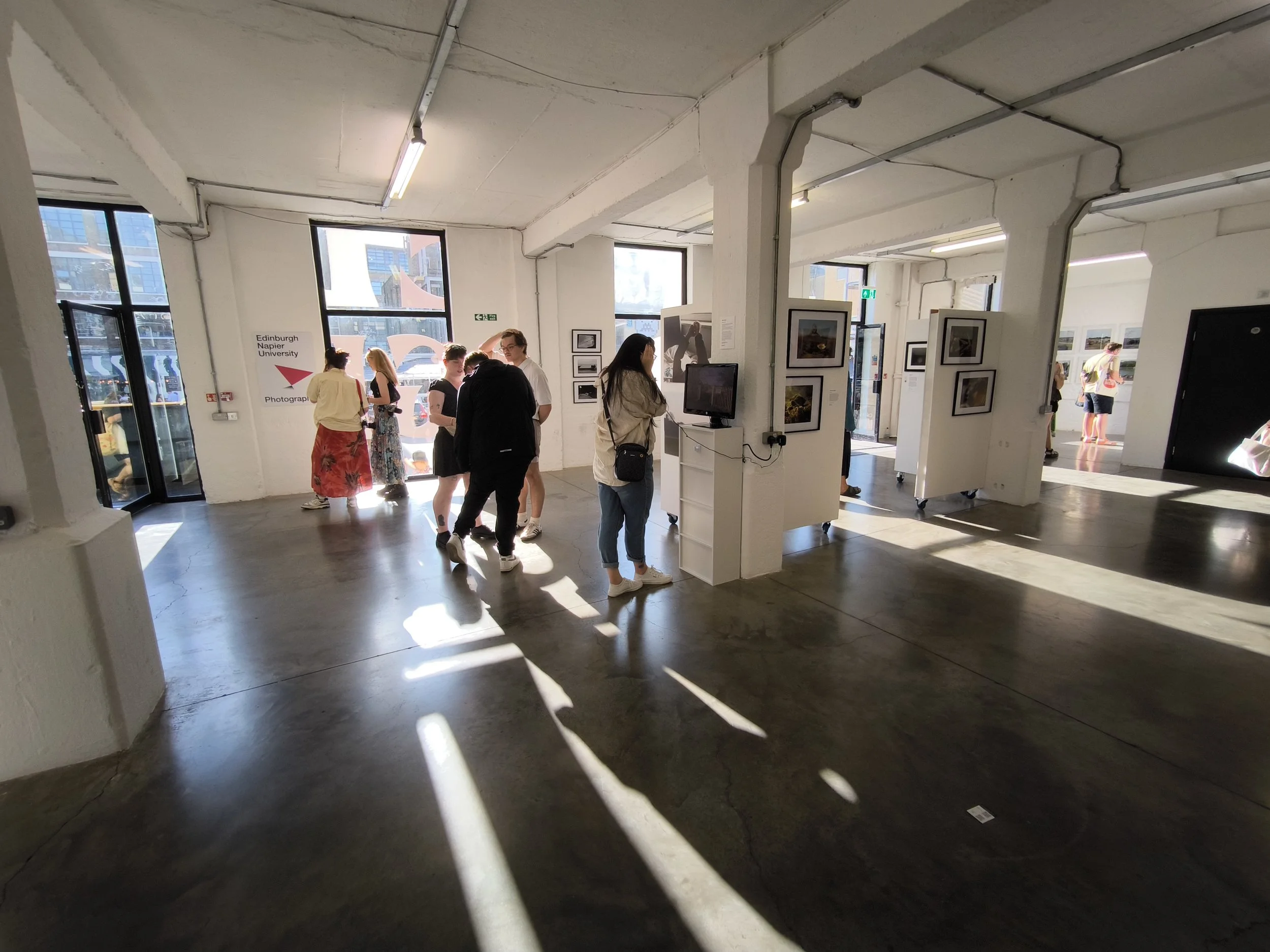 People inside an art gallery with photographs on the walls, lit by natural light streaming through large windows.