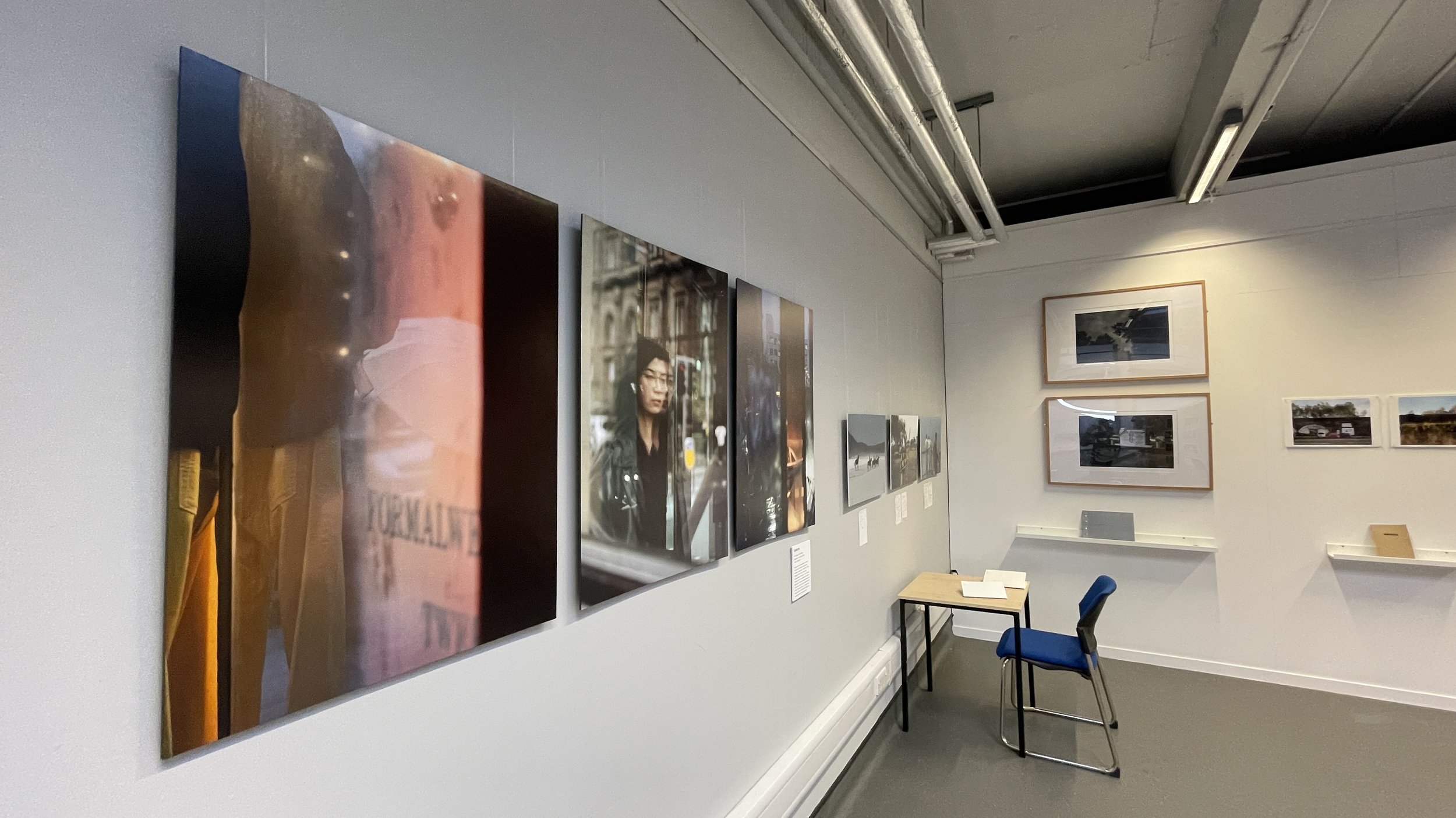 Photo of an art gallery with various photographs displayed on the walls, a small table with a book and a blue chair, and two large framed photographs on the wall in the background.