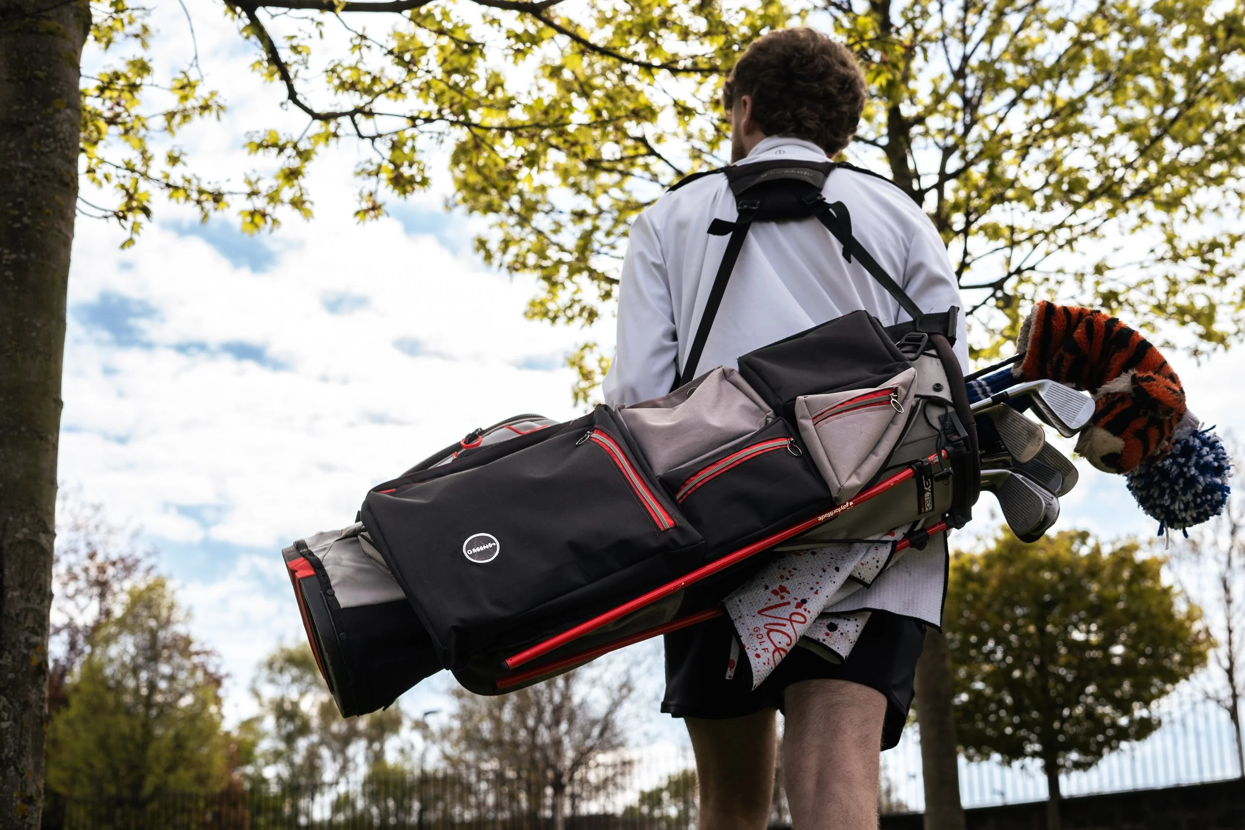 A young man in golf attire walking on a golf course through an area with trees, away from the camera with a golf bag on his back