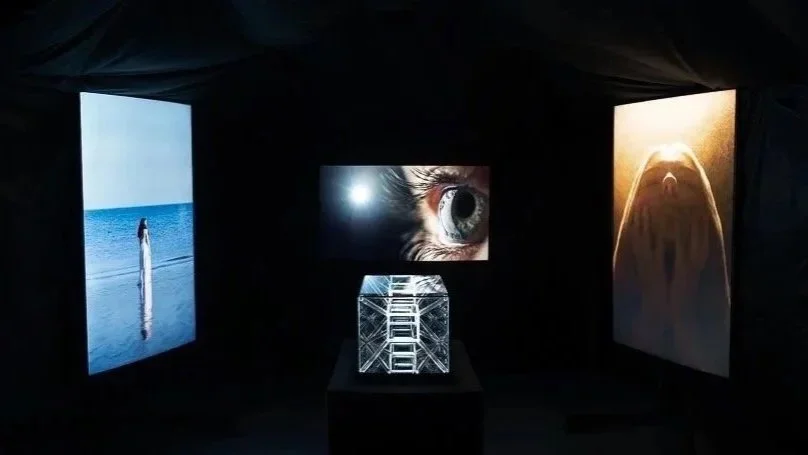 An art exhibition with three large digital screens displaying images: a woman in white on the beach, a close-up of a human eye, and a figures in silhouette with a beard. There is an illuminated glass box with the text "FUTURE" in the center.