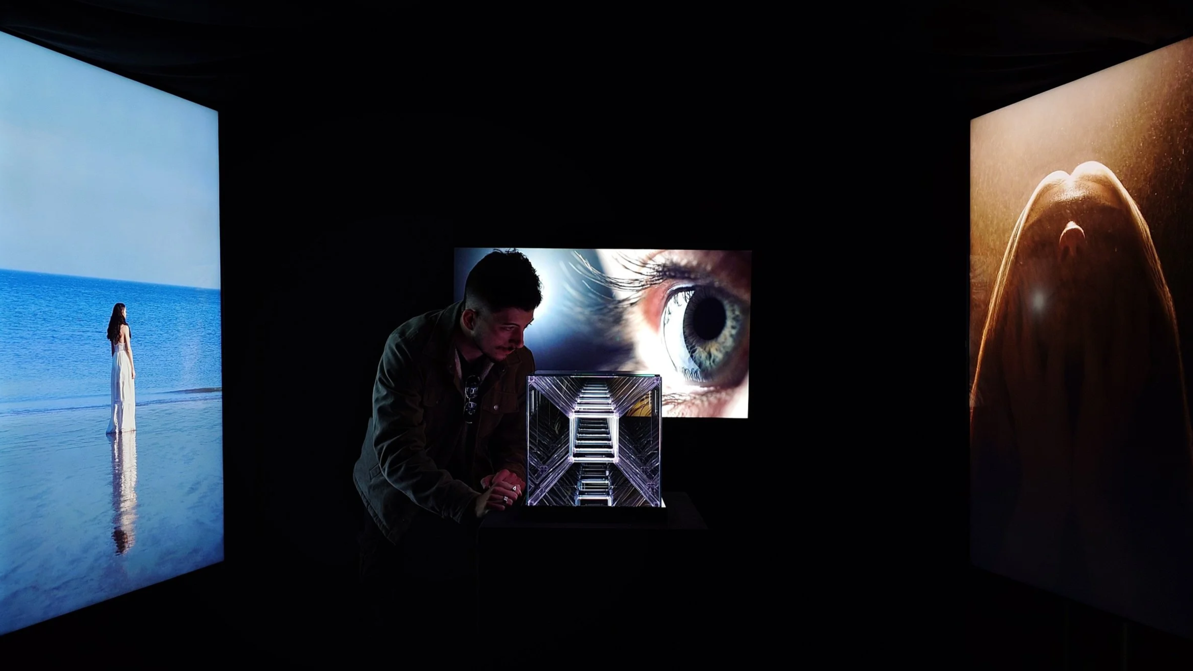 A person observing digital art displays in a dark gallery, including a large eye close-up, a woman by the ocean, and a mountainous landscape.