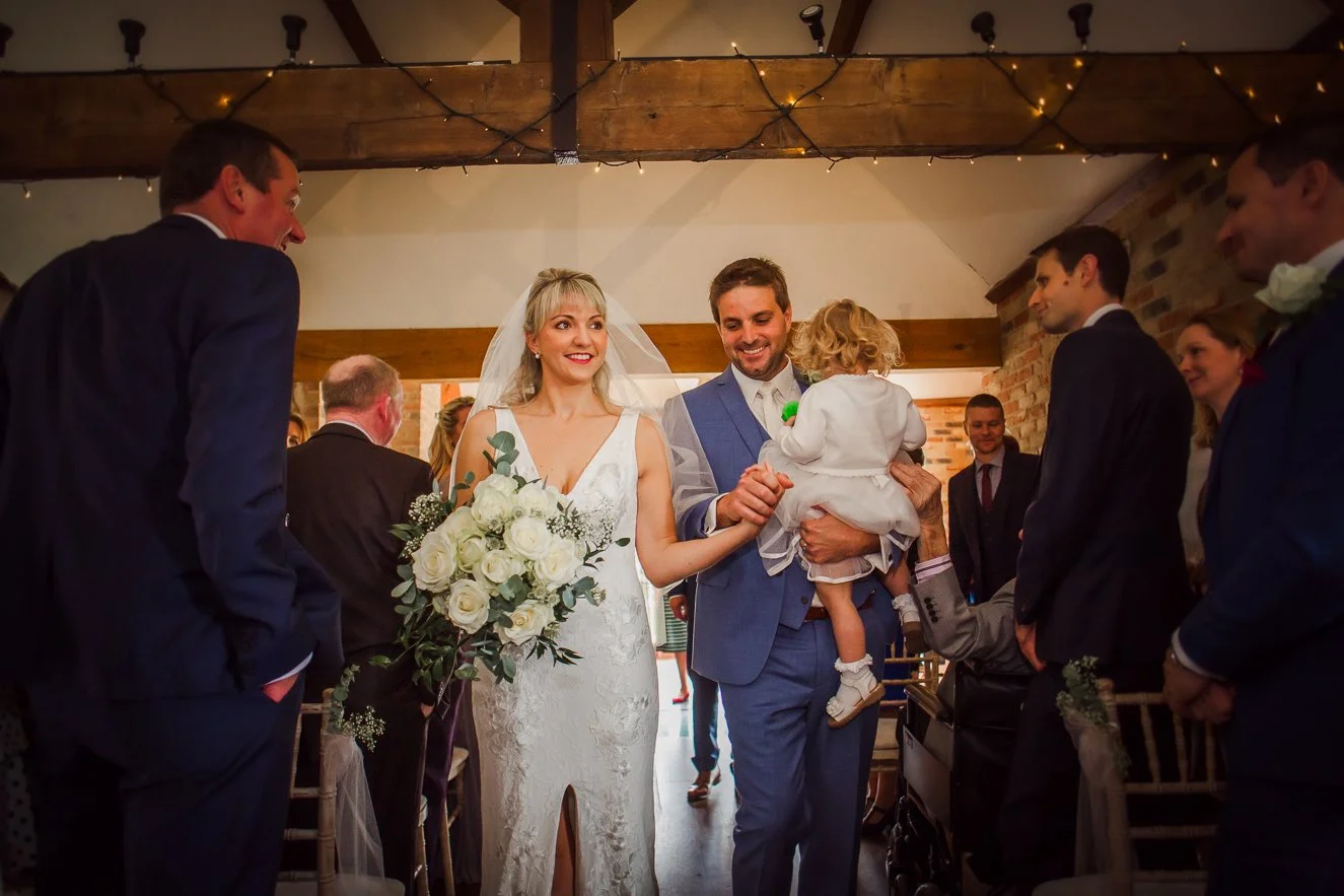 Bride and groom walking down the aisle at their wedding reception, surrounded by friends and family in a rustic venue with brick walls and wooden beams.