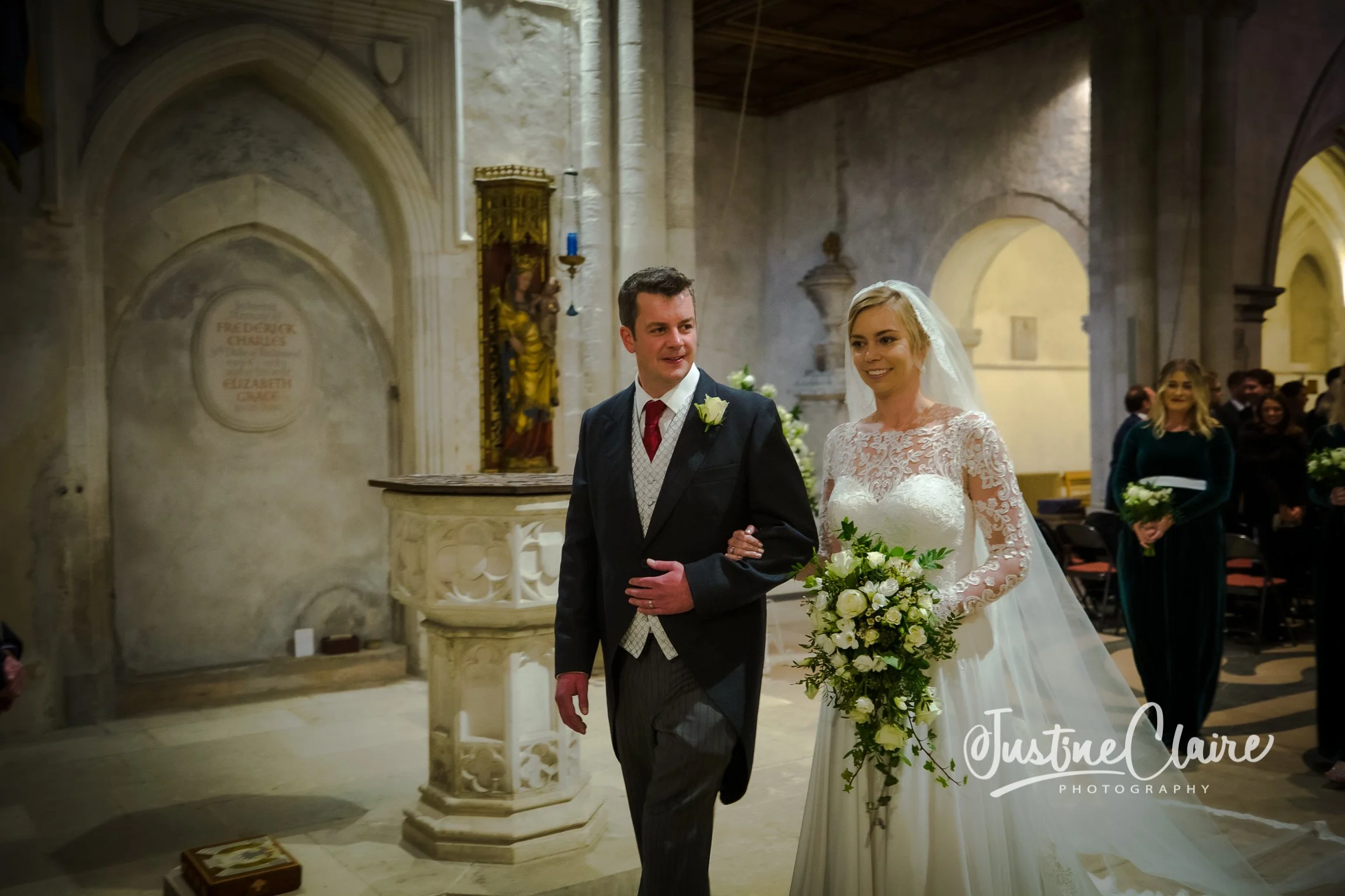 A bride and groom walking down the aisle inside a church, with the bride holding a bouquet of white flowers and the groom in a black suit. Bridesmaids and guests are visible in the background.