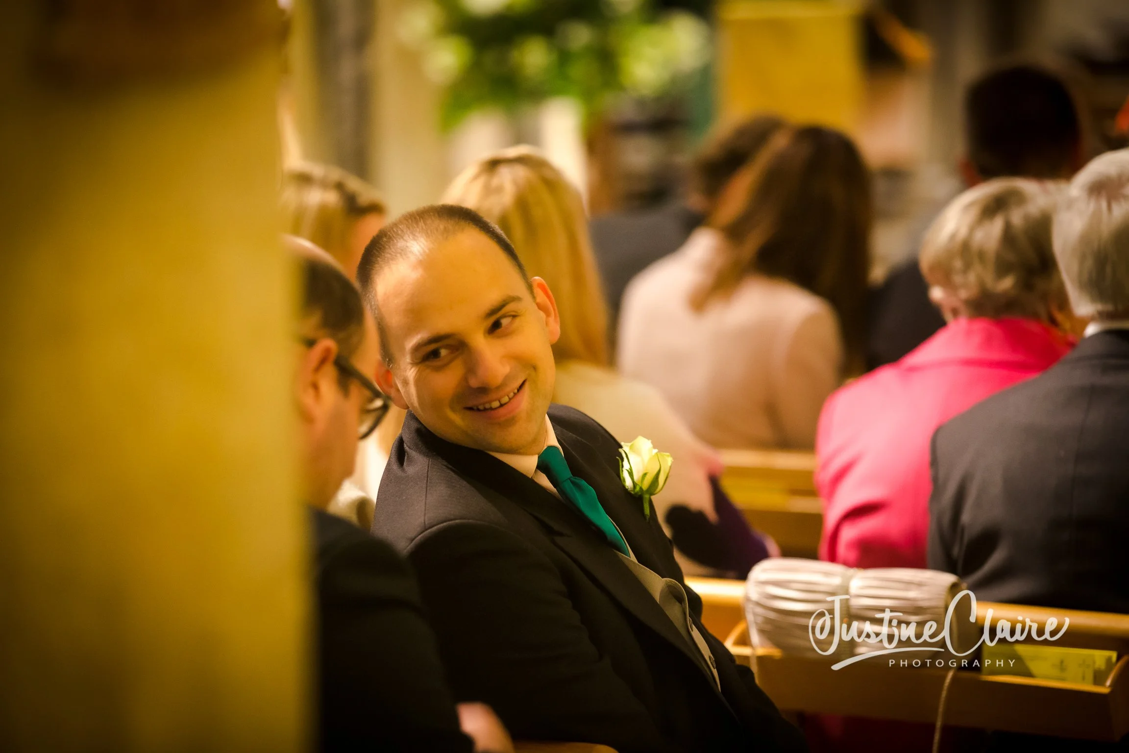 A man in a dark suit with a white shirt and green tie, smiling and looking to his left, sitting among a group of people in a gathering, likely a wedding or formal event, inside a warmly lit venue.