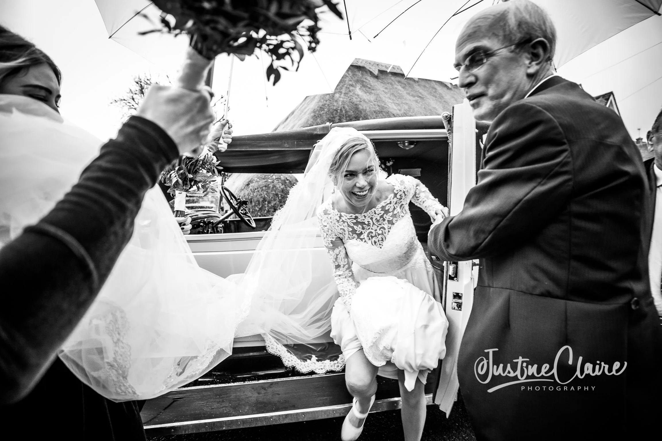 A bride smiling as she steps out of a vehicle on her wedding day, assisted by a man in a suit. A person holding an umbrella is partially visible in the foreground. The scene appears to be outdoors with a house in the background.