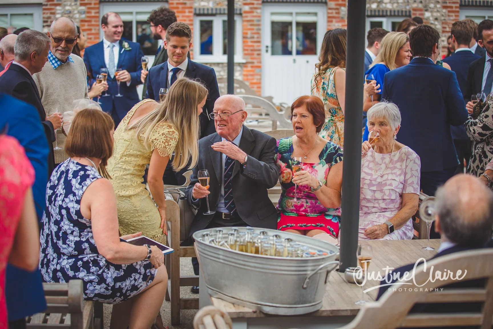 Group of mostly senior adults socializing at an outdoor gathering, some sitting and some standing, holding glasses of champagne, with a brick building in the background.