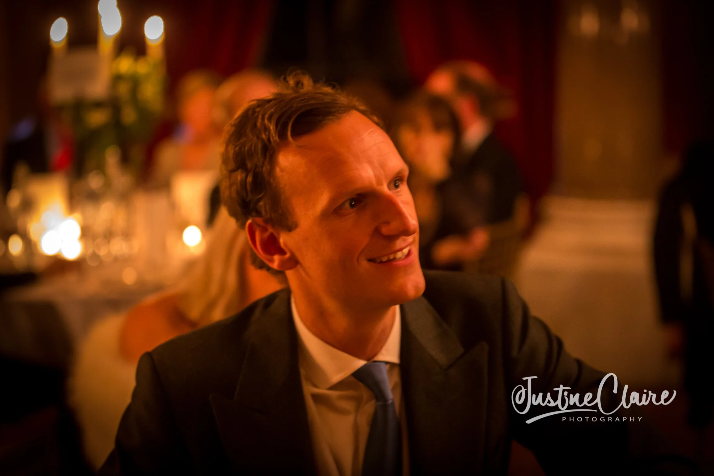A man in a suit and tie smiling at an indoor event or banquet, with warm lighting and blurred background of other guests.