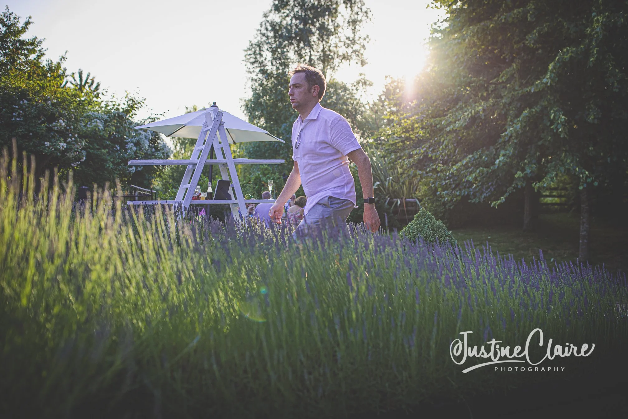 Man walking through a lavender field at sunset with trees and a white outdoor bar in the background.
