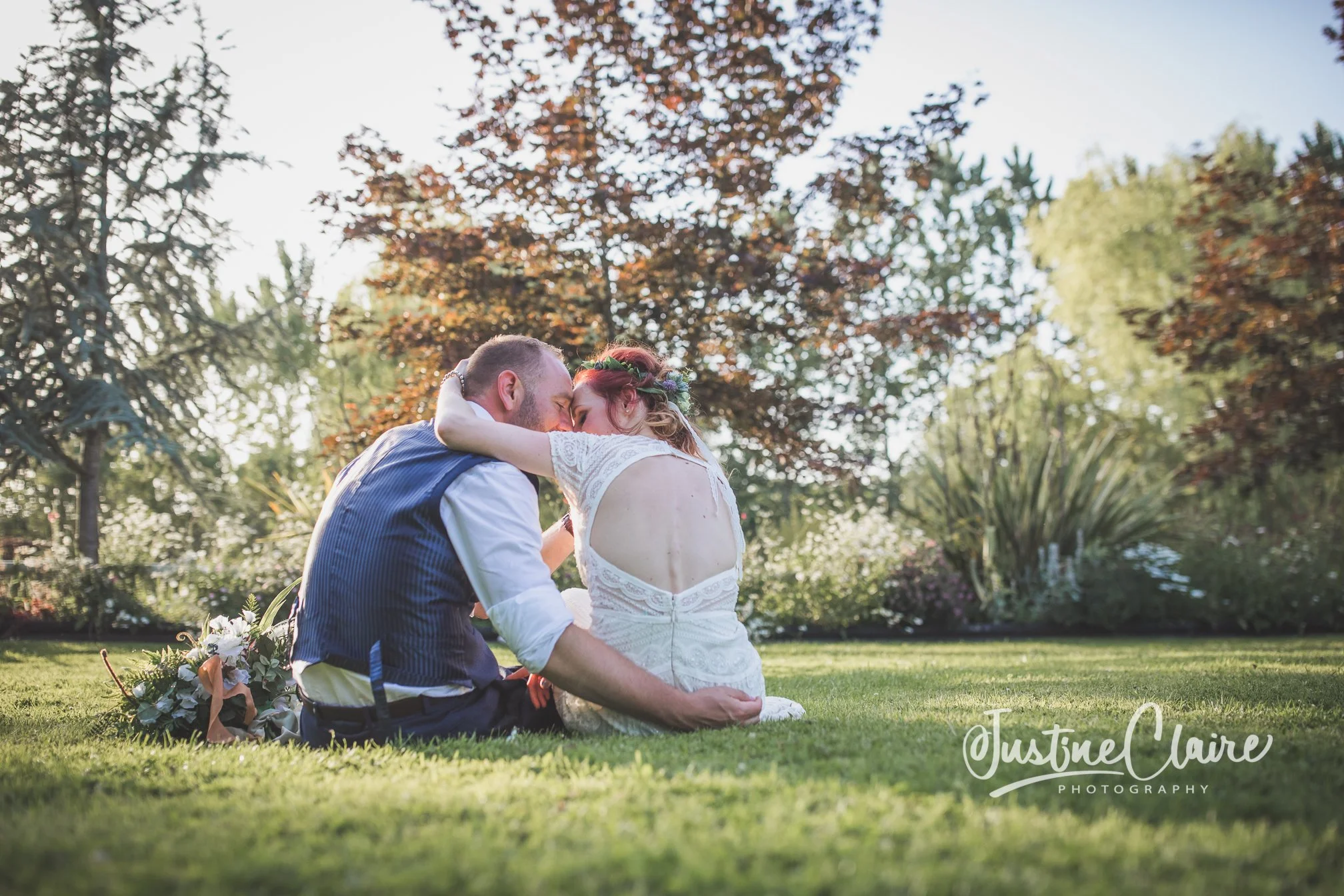 A newlywed couple sits on grass in a park, embracing and touching foreheads, with trees and sunlight in the background.