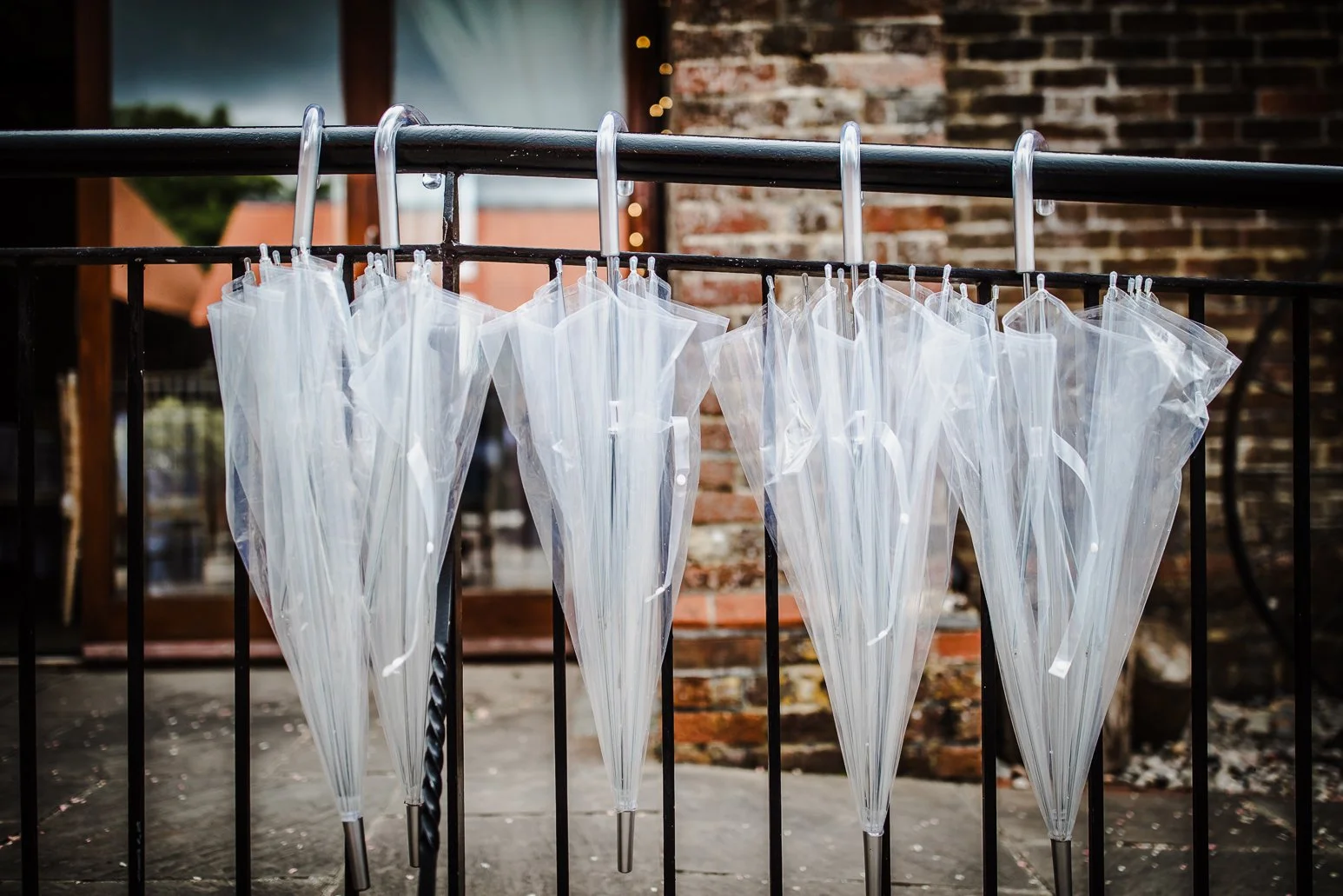 Multiple clear umbrellas hanging upside down on a black metal railing outside with a brick building in the background.