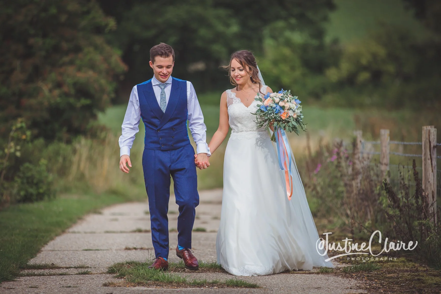 A couple in wedding attire walking hand in hand on a rural path, with trees in the background. The bride is holding a bouquet of flowers and wearing a white gown, while the groom is dressed in a blue vest and pants with a light blue shirt. They are s