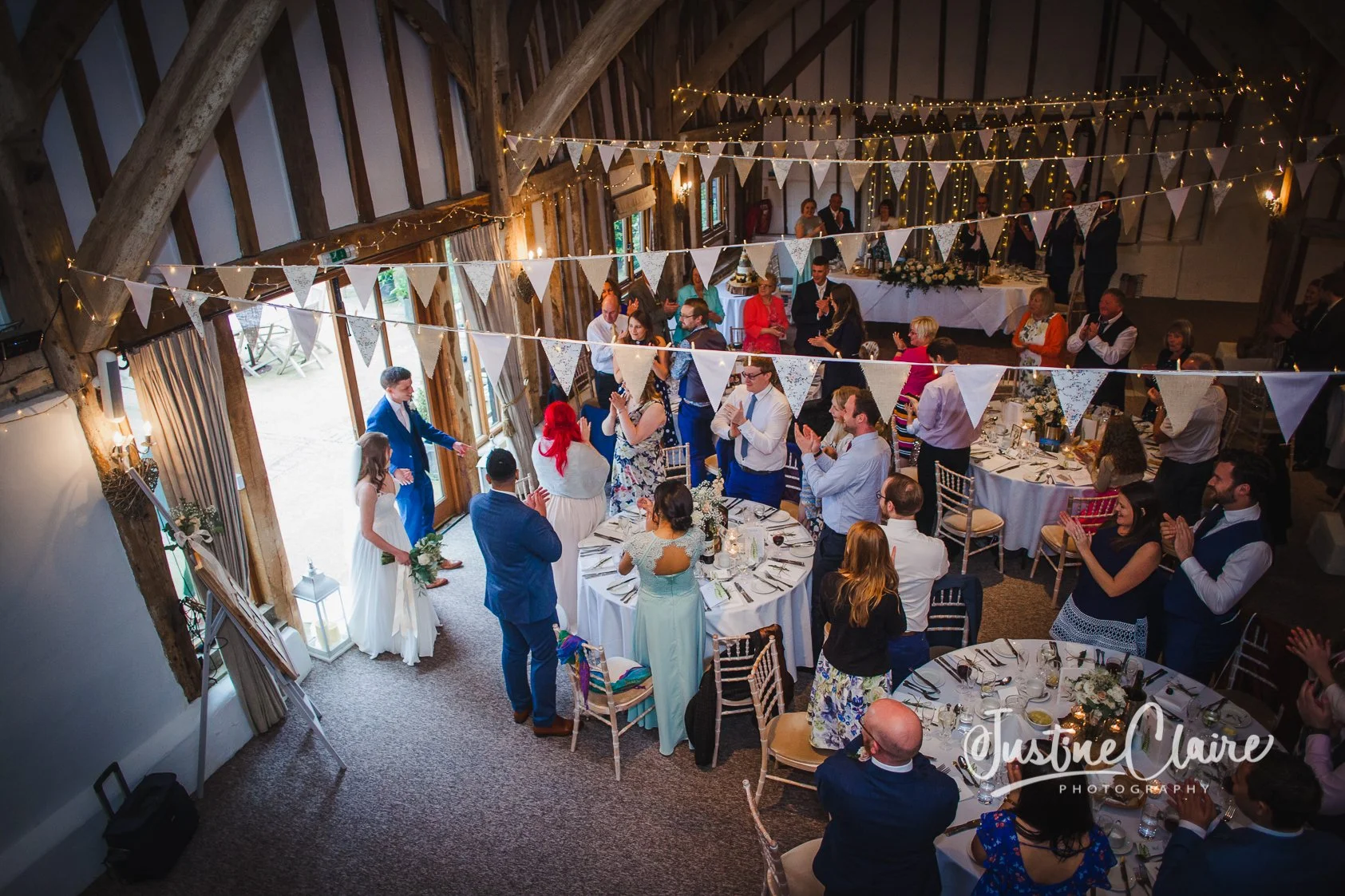 Wedding reception with guests clapping and smiling inside a rustic barn decorated with string lights and white bunting banners.