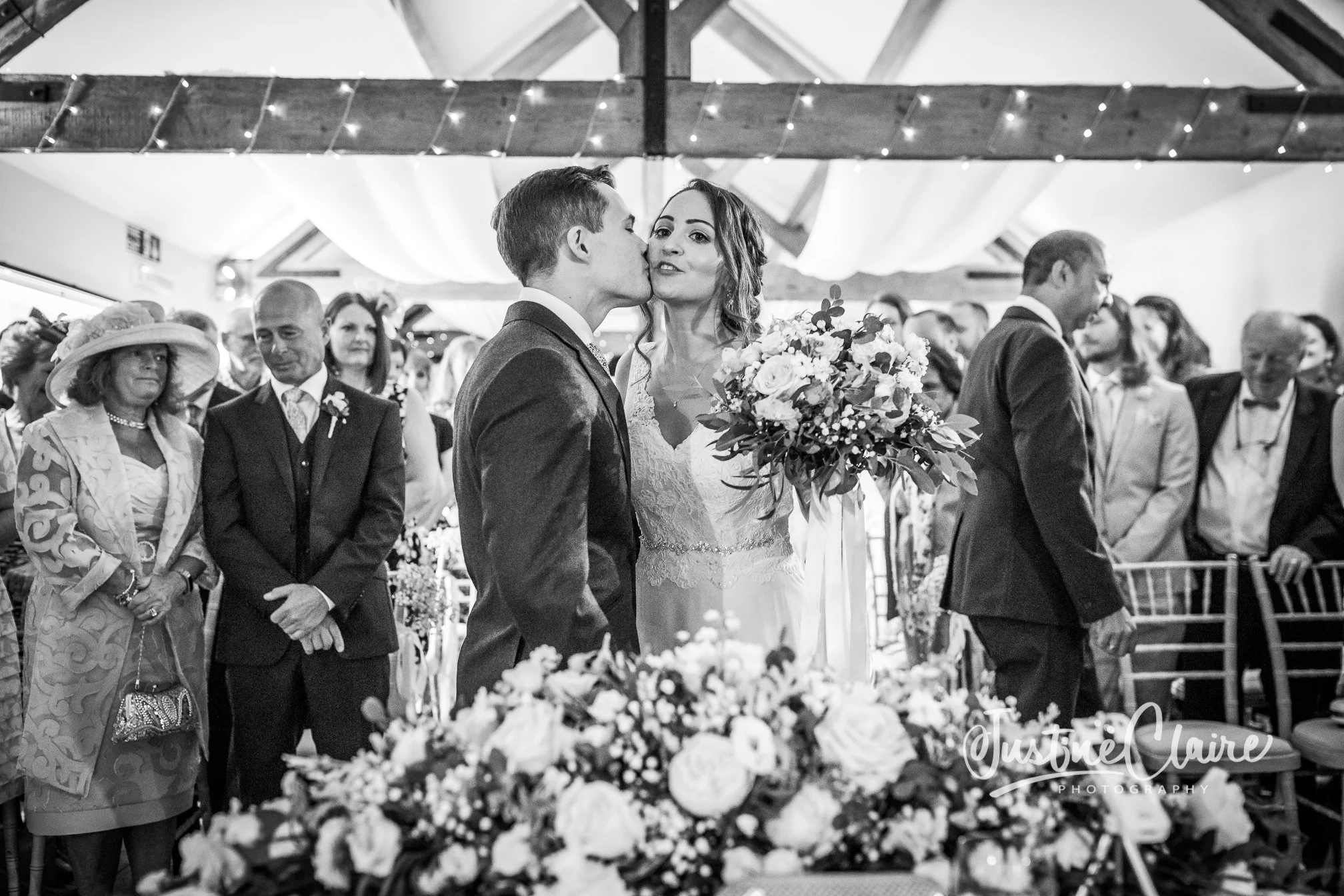 A black and white photo of a wedding ceremony with the bride and groom at the center. The groom is kissing the bride on the cheek while she holds a bouquet of flowers. Guests are standing and watching in the background inside a decorated venue.