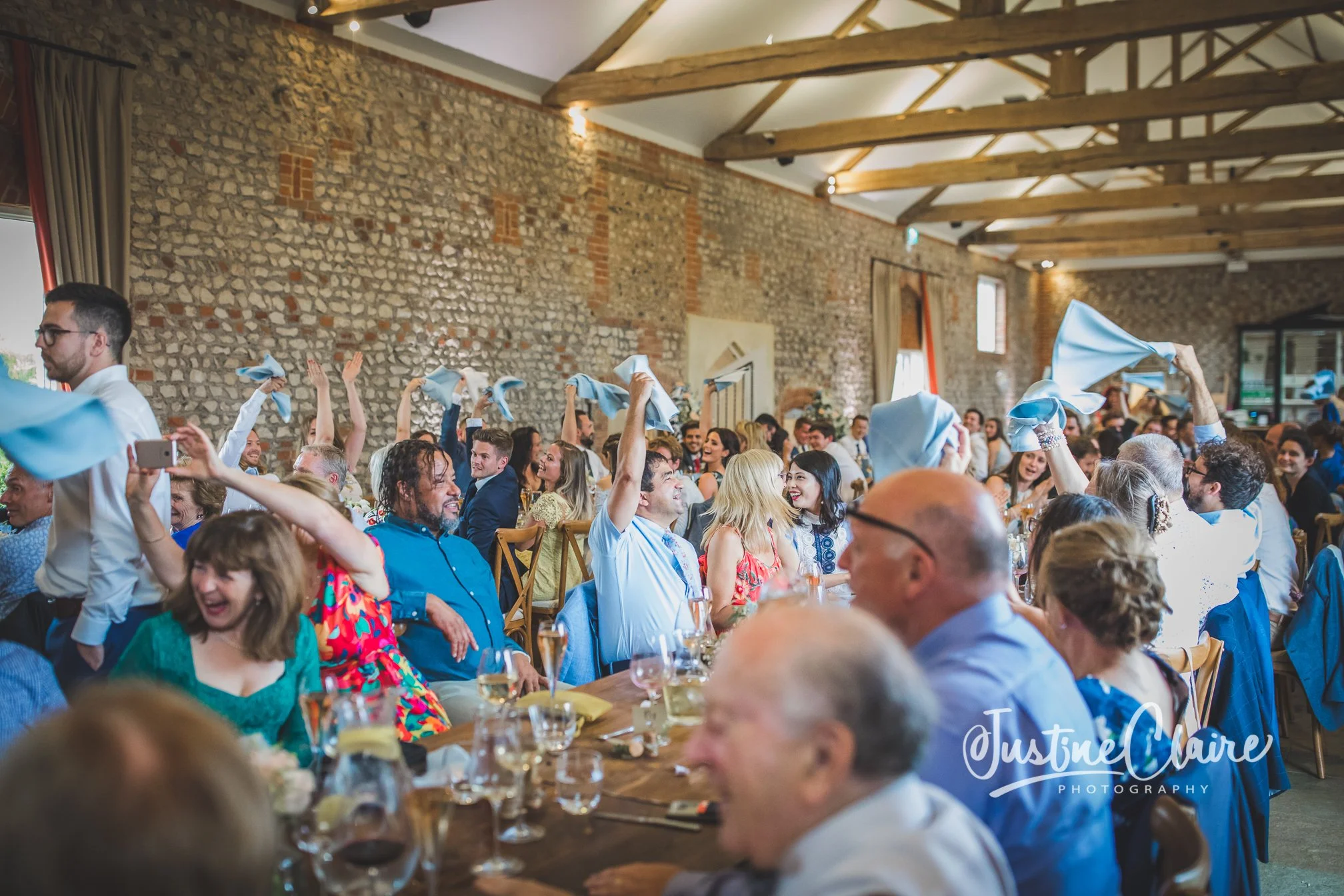 Guests at a celebration or wedding reception dancing and waving light blue napkins in a rustic venue with brick walls and wooden beams.