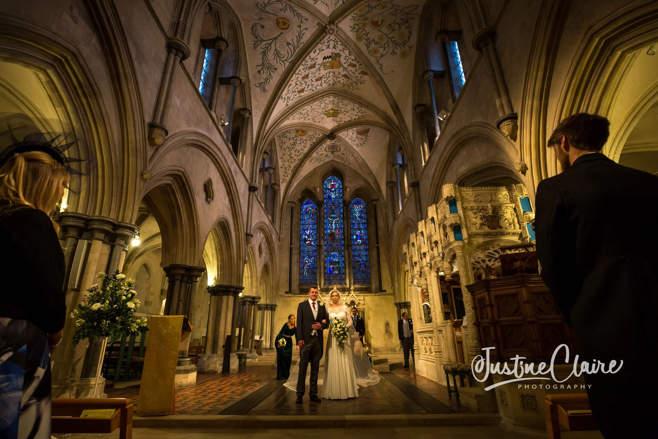 A wedding ceremony inside a church with stained glass windows and detailed architectural features. The bride and groom stand at the altar, surrounded by guests. Boxgrove Priory wedding photographers