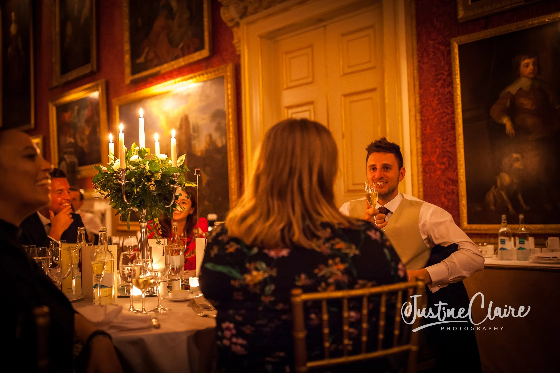 People celebrating at a formal dinner, with a man holding a glass of champagne smiling, in a decorated room with paintings and a candelabra at Goodwood House