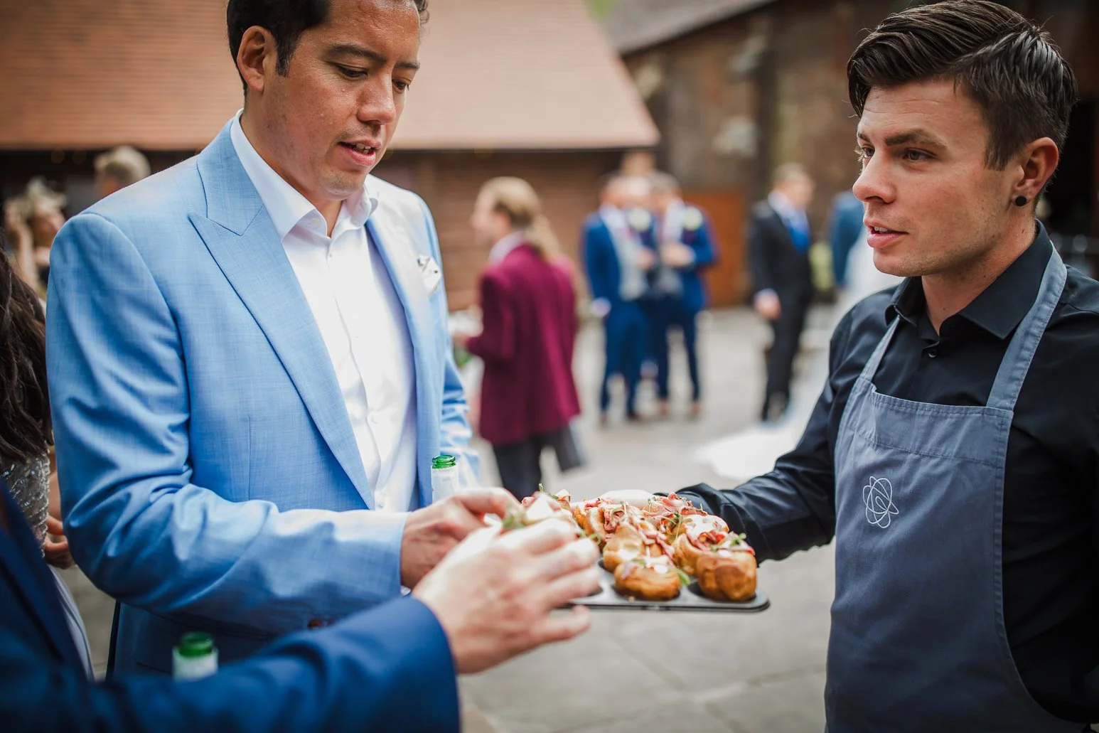 Man in a blue suit receiving appetizers from a man in a black shirt and apron at an outdoor event.