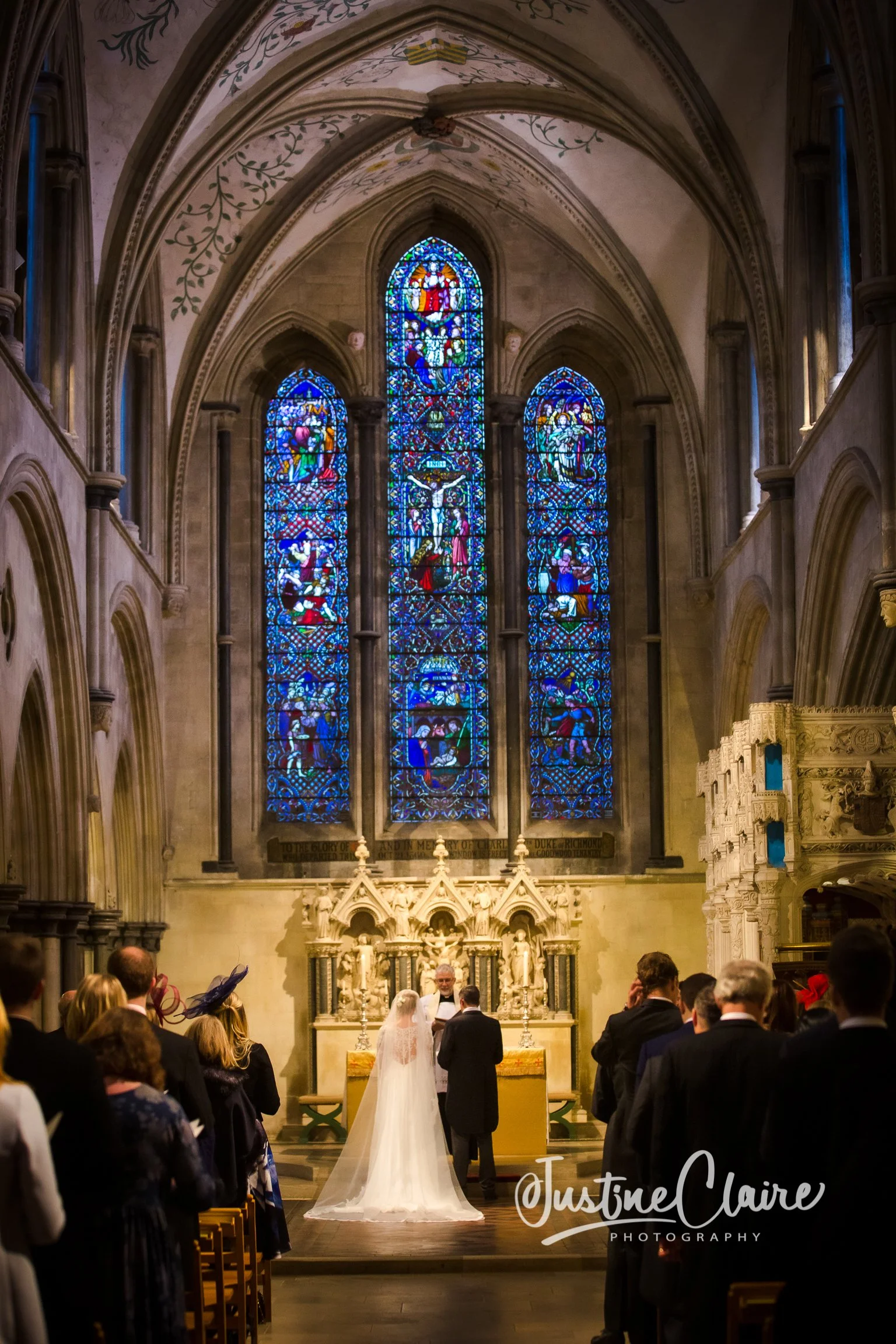 A wedding ceremony inside a church with stained glass windows, a bride and groom standing before a minister, and guests seated and standing around.