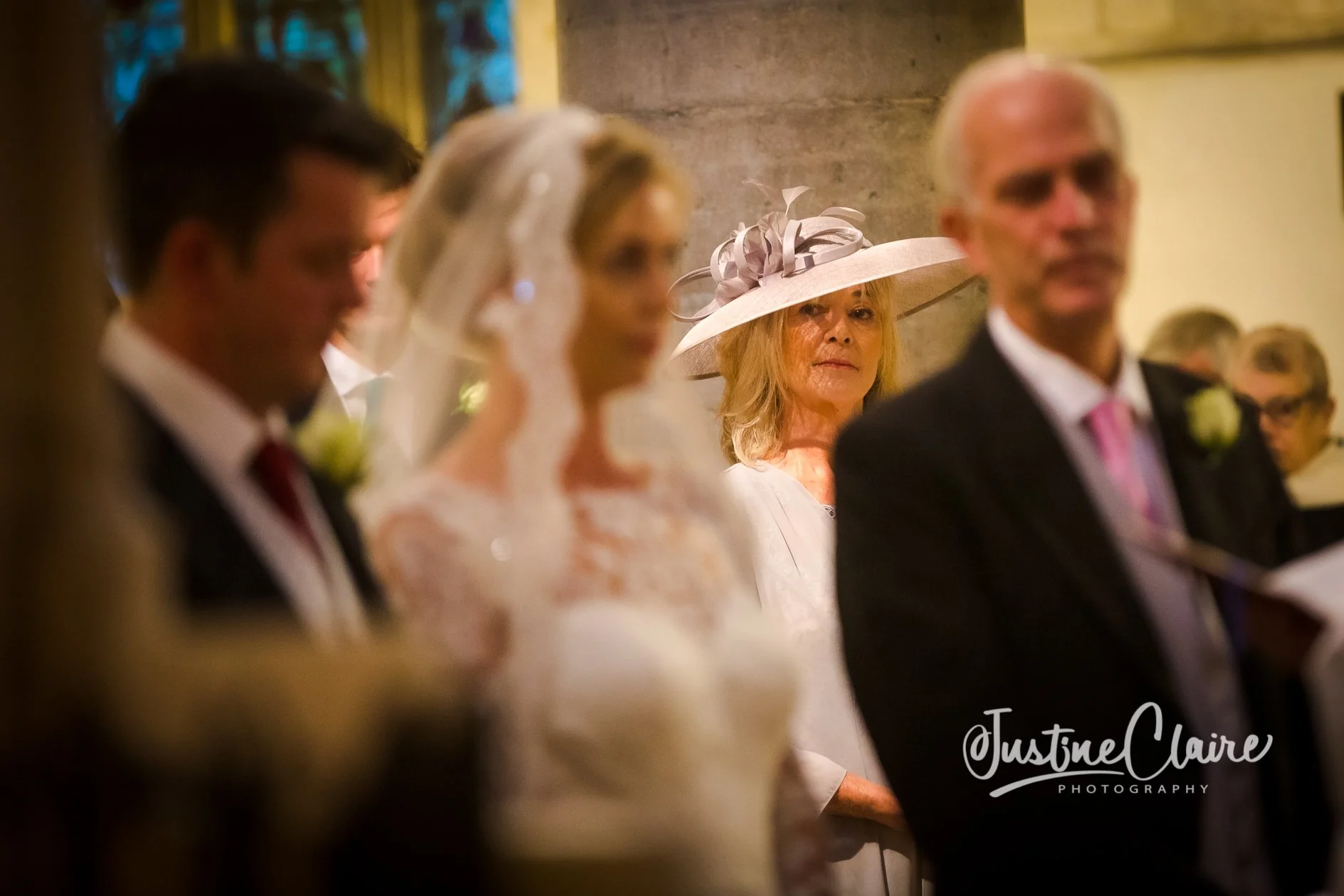 A woman in a large, elegant hat with ribbons at a wedding, focused on by others including a man and woman in formal attire and glasses, in a church setting.