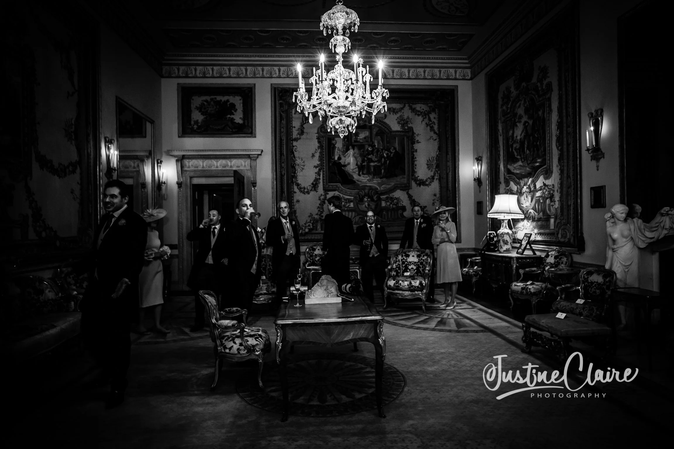 A group of well-dressed people, mostly men in suits, gathered in an elegant, richly decorated room with ornate furniture, artwork, and a large chandelier, appearing to be at a formal social event or gathering.