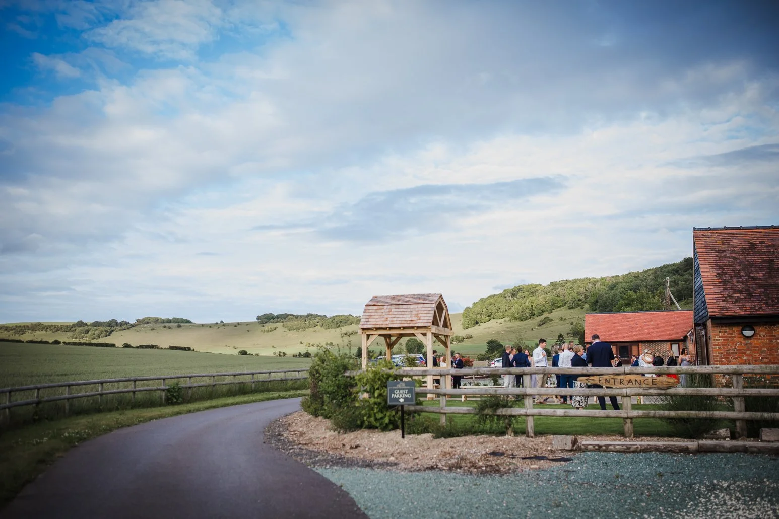 Guests gathering outside a rustic venue with buildings made of red bricks, a wooden entrance sign, and a scenic countryside landscape in the background under a partly cloudy sky.