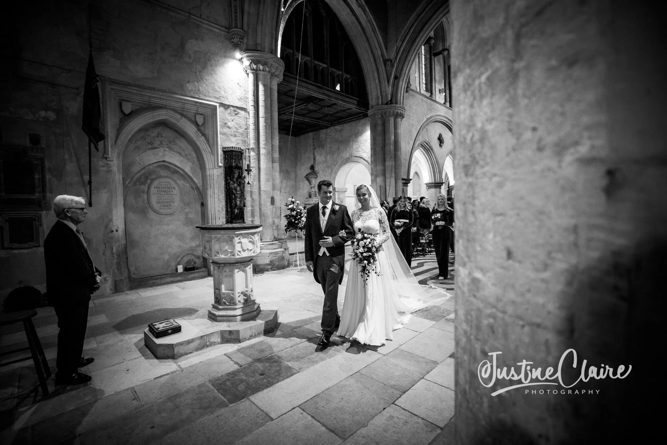 Black and white photo of a bride and groom walking down the aisle in a church, with guests in the background and an officiant on the left