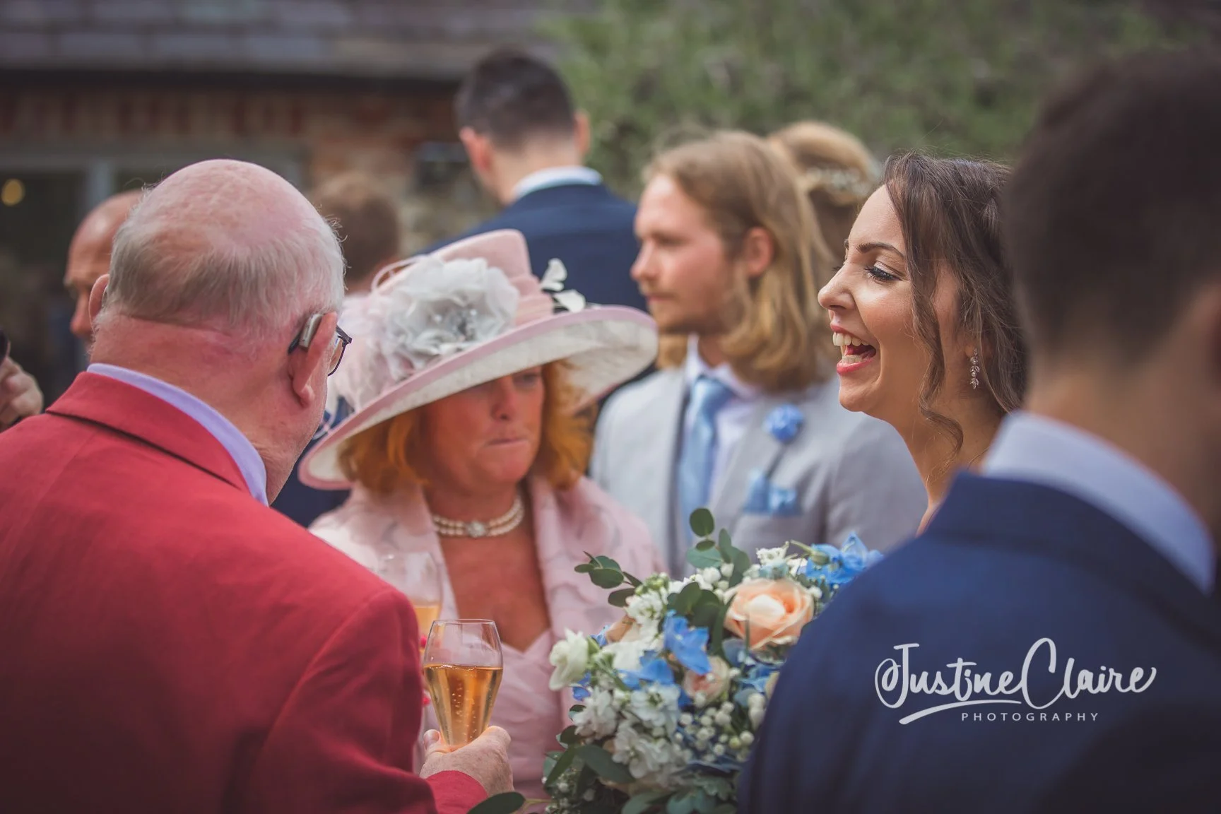 Group of people at a wedding reception, including a woman in a light gray suit holding a bouquet and a woman in a pink dress with a large pink hat, exchanging words with a man in a red jacket holding a glass of champagne. candid wedding photographer