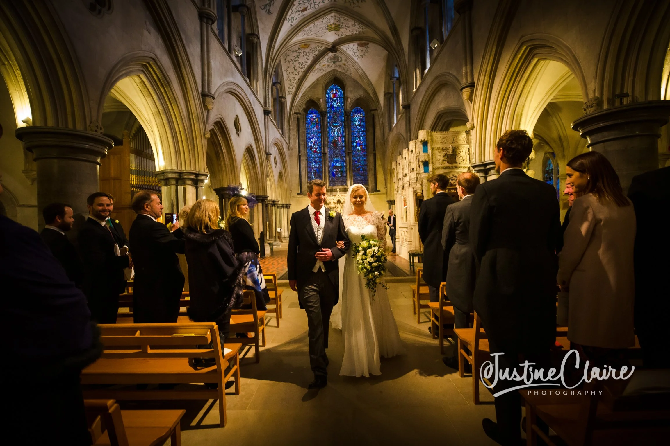 A bride and groom walk down the aisle, smiling, inside a historic church with stained glass windows. Guests are standing and taking photos, with warm lighting and tall stone arches.