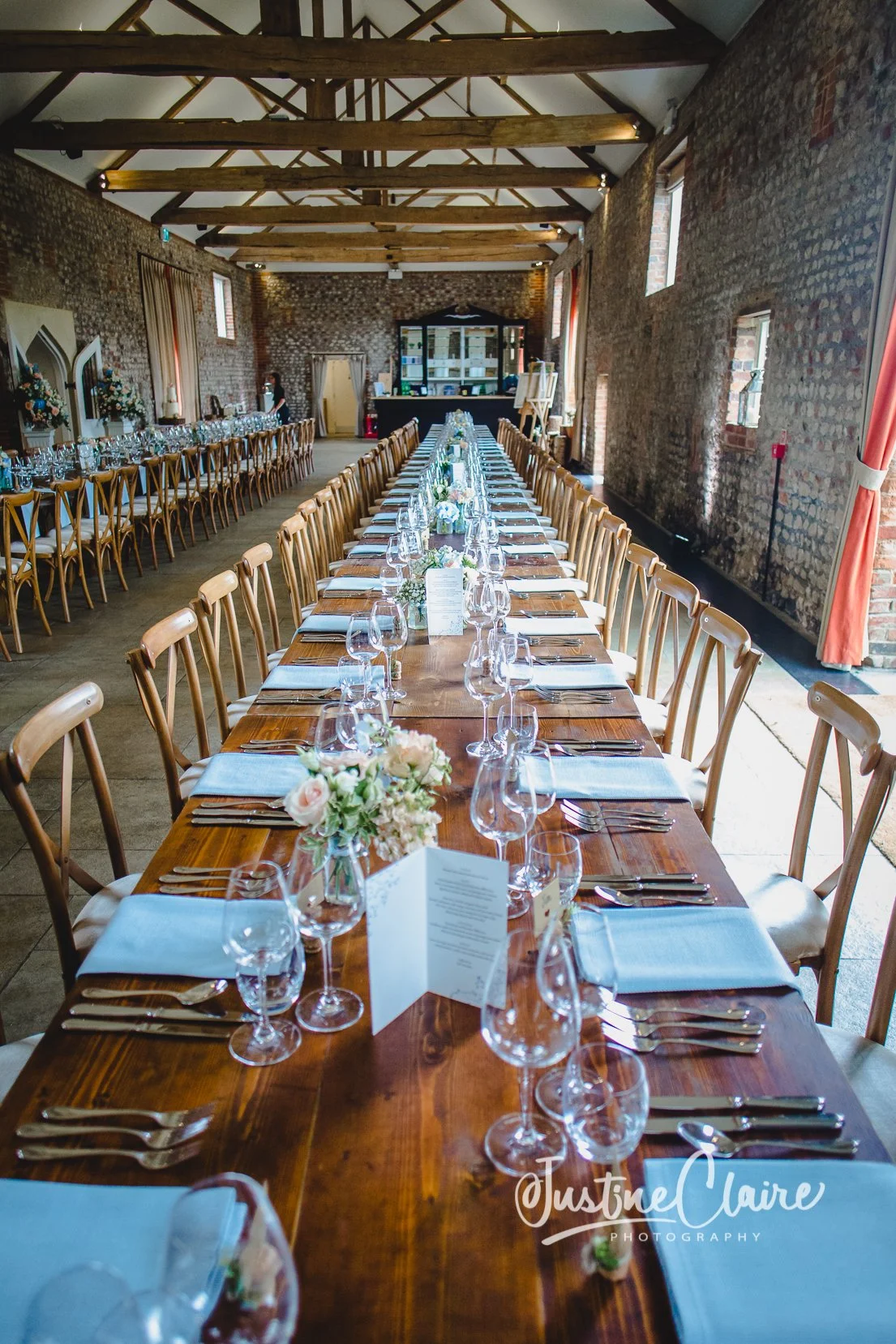 Long wooden banquet table set for a wedding reception with white flowers, glassware, and silverware inside a rustic brick venue with high ceiling beams.