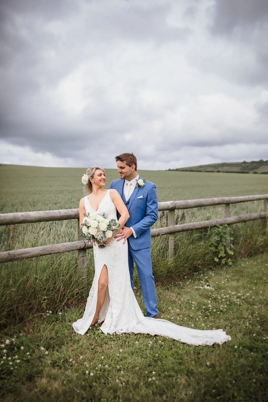 A bride in a white gown holding a bouquet of white roses and greenery, standing beside a groom in a blue suit, outdoor setting with a grassy field and cloudy sky.