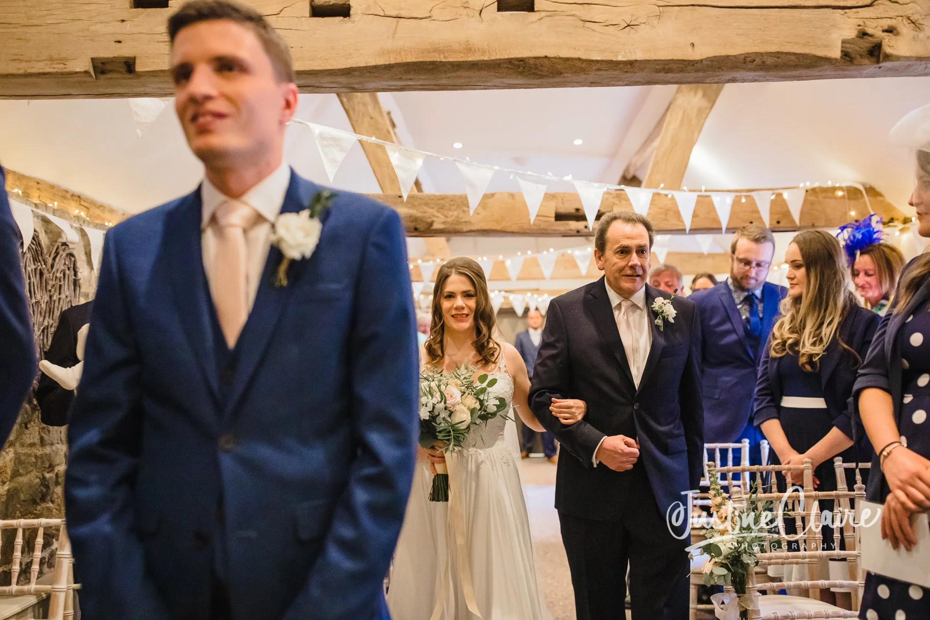 People at a wedding ceremony, with a bride holding a bouquet walking behind a man in a dark suit, as others stand in the background inside a decorated venue with bunting and string lights ceremony at Fitzleroi barn