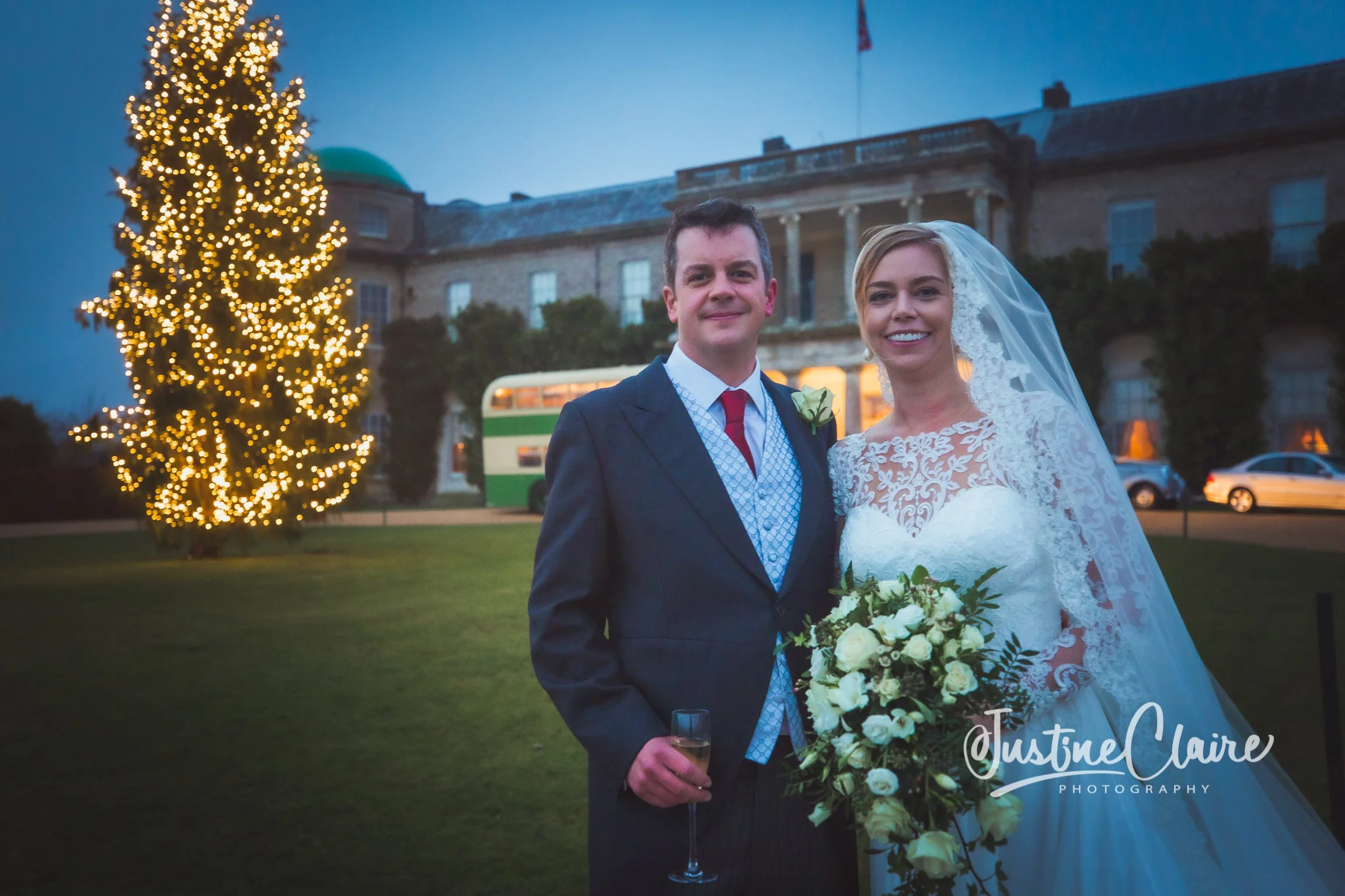 A newlywed couple smiling outside at dusk, with a decorated Christmas tree and a historic building in the background.
