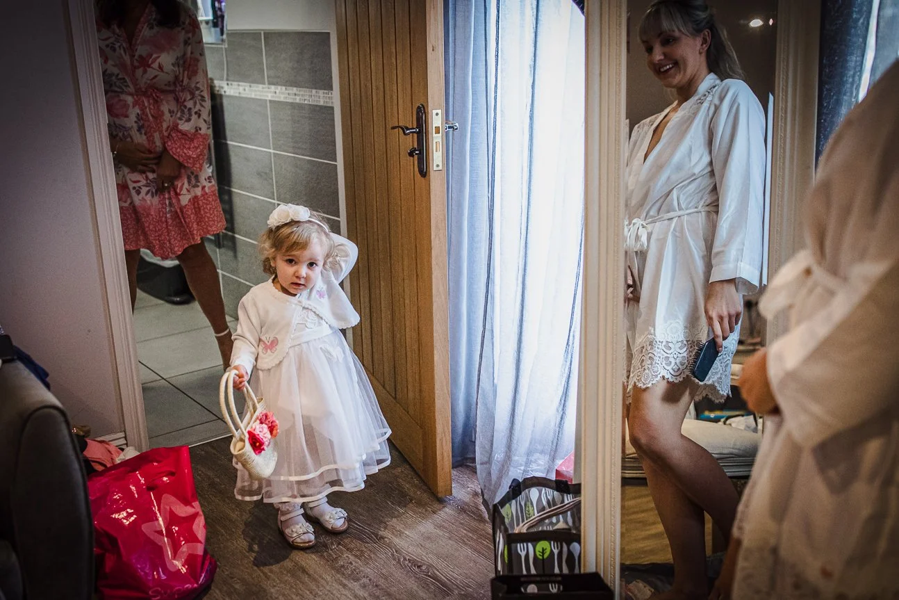 A young girl in a white dress and bows is holding a small basket with flowers, standing at a doorway. Two women, also in white robes, are visible; one is smiling and the other is partially visible. The girl appears to be about to go through the door,