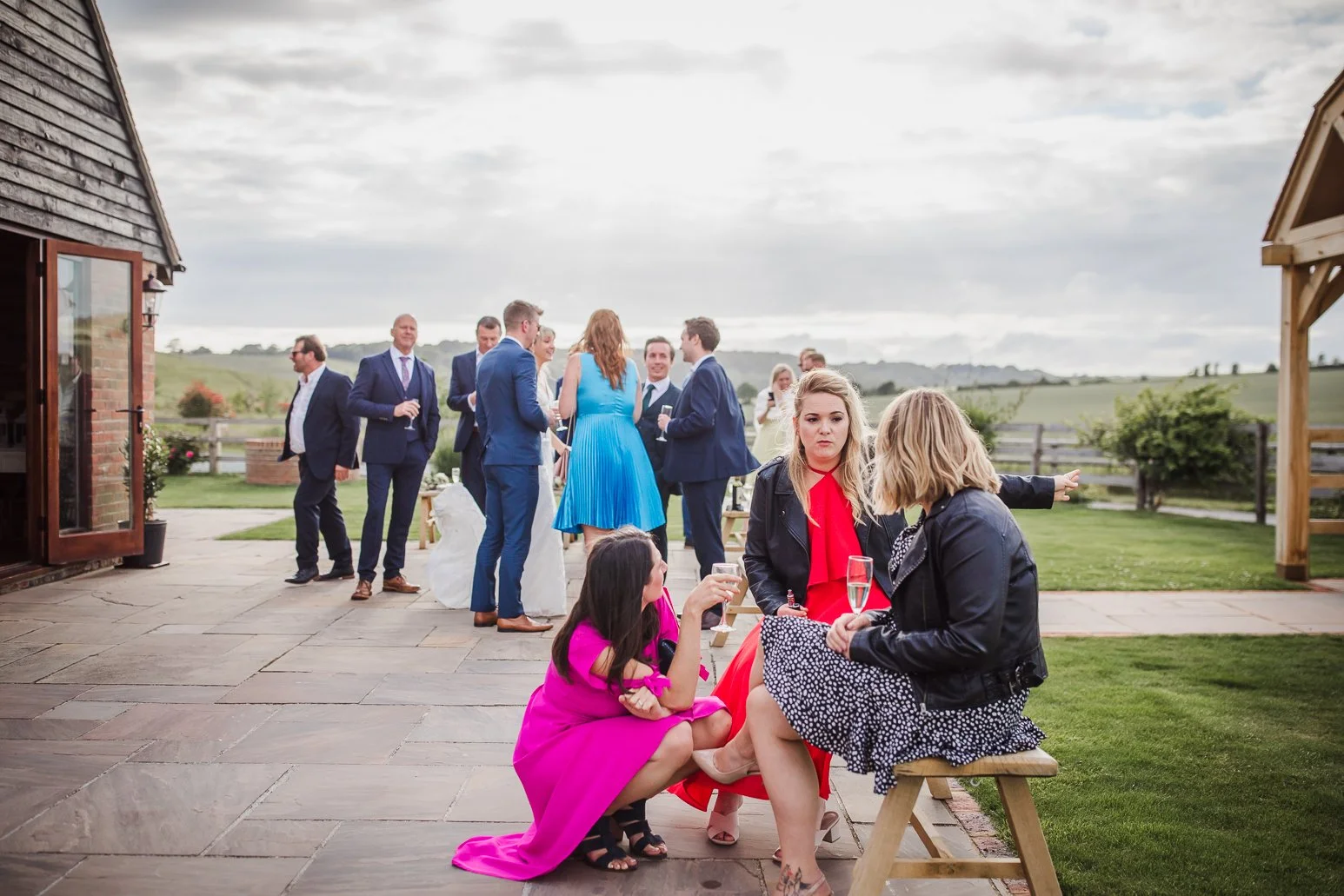 People at an outdoor social gathering, some standing and chatting, others sitting on a bench, with a rural landscape in the background.